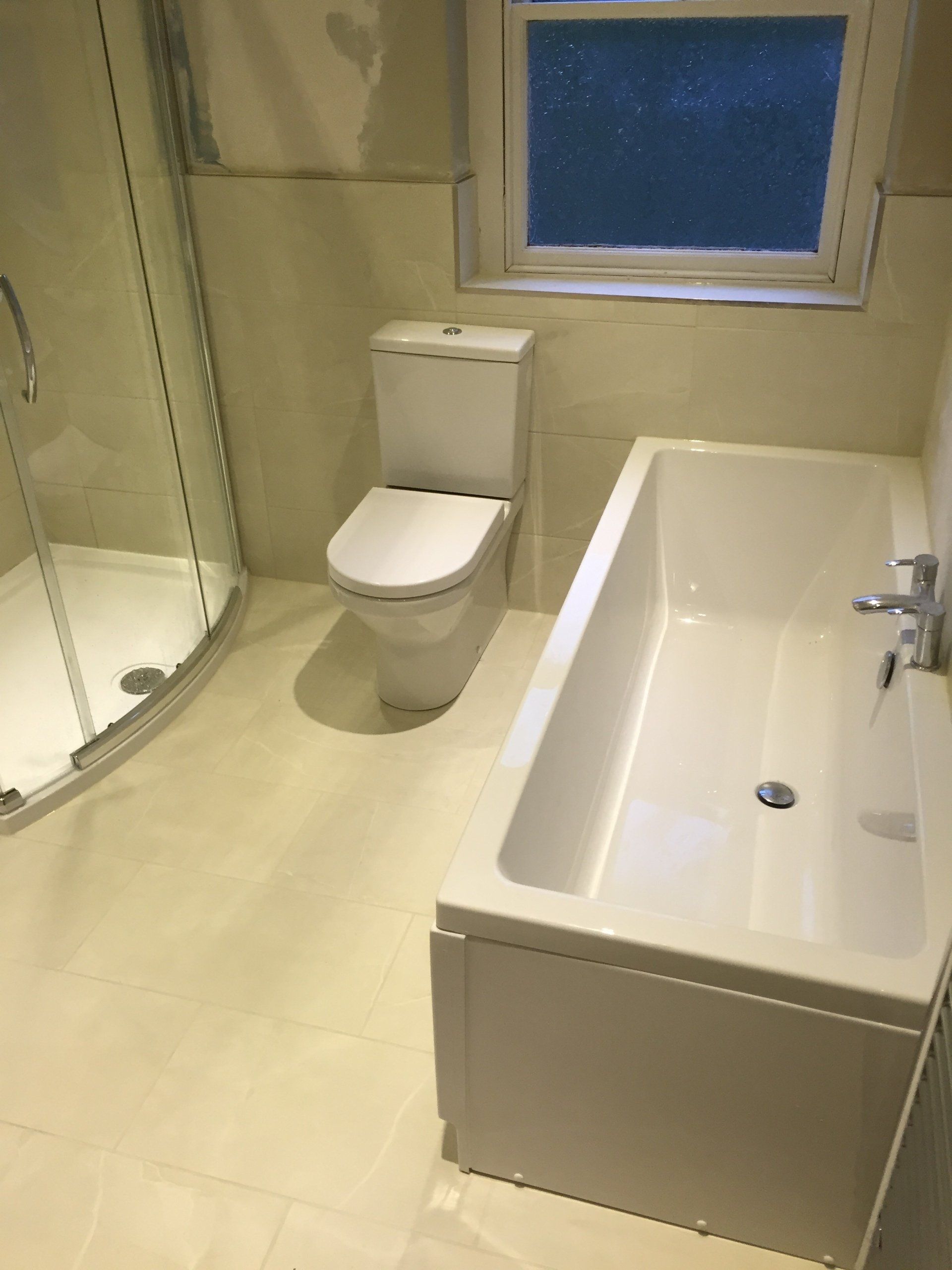 Bathroom with shower, toilet, and bathtub; beige tile; window above the tub.