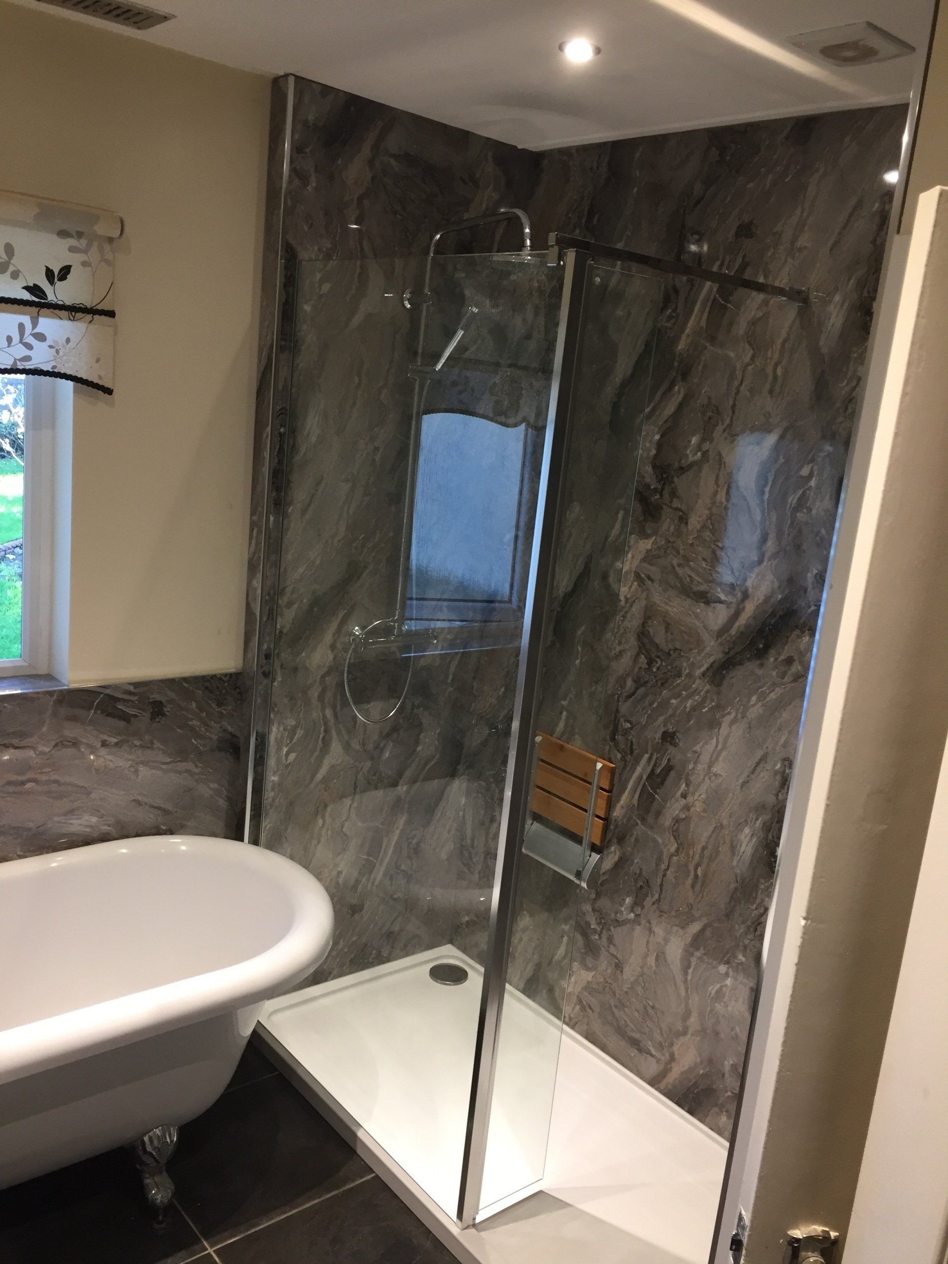 Bathroom with a grey marble-effect shower enclosure, a white tub, and a window with a floral blind.