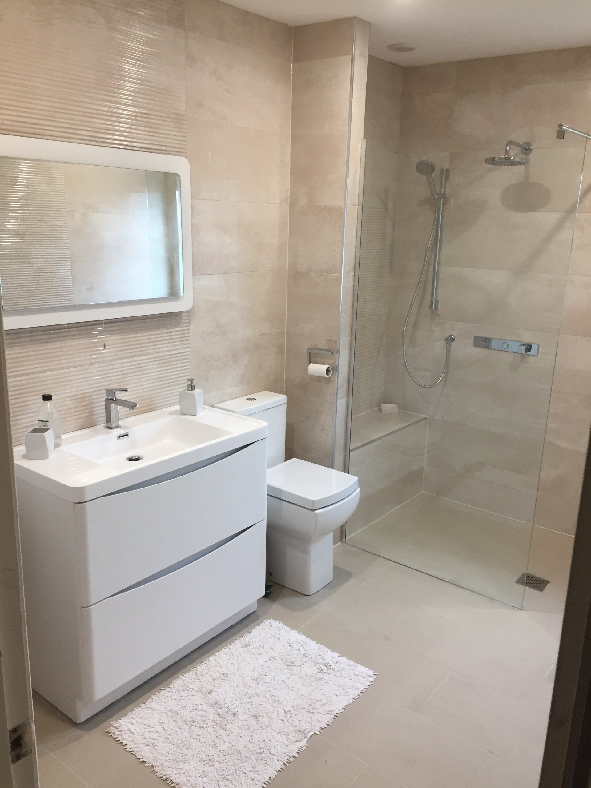 Modern bathroom with a white vanity, toilet, and shower. Neutral beige tiles and a white bath mat.