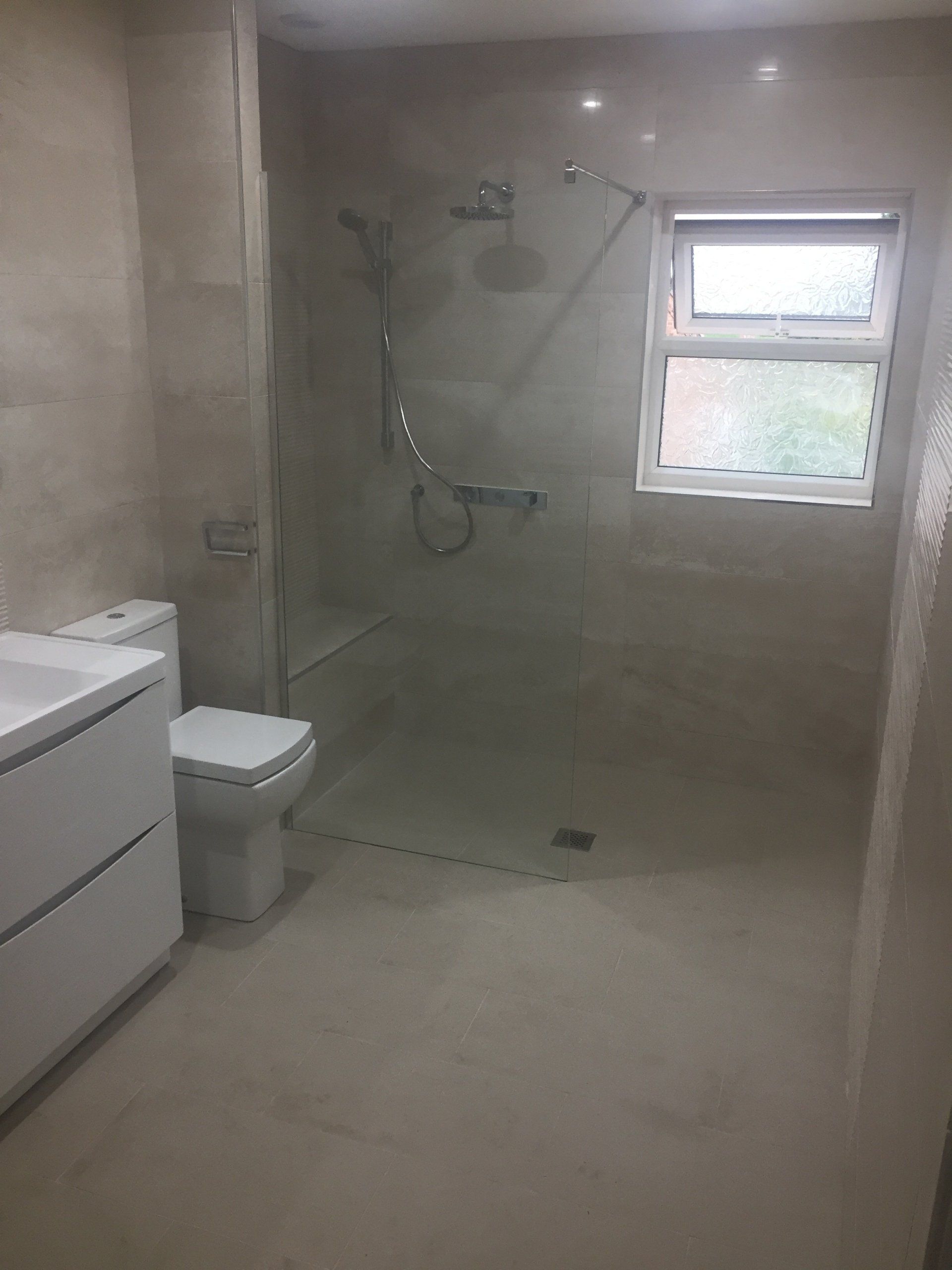 Modern bathroom with a glass shower enclosure, toilet, vanity, and window. Light beige tiles and walls.