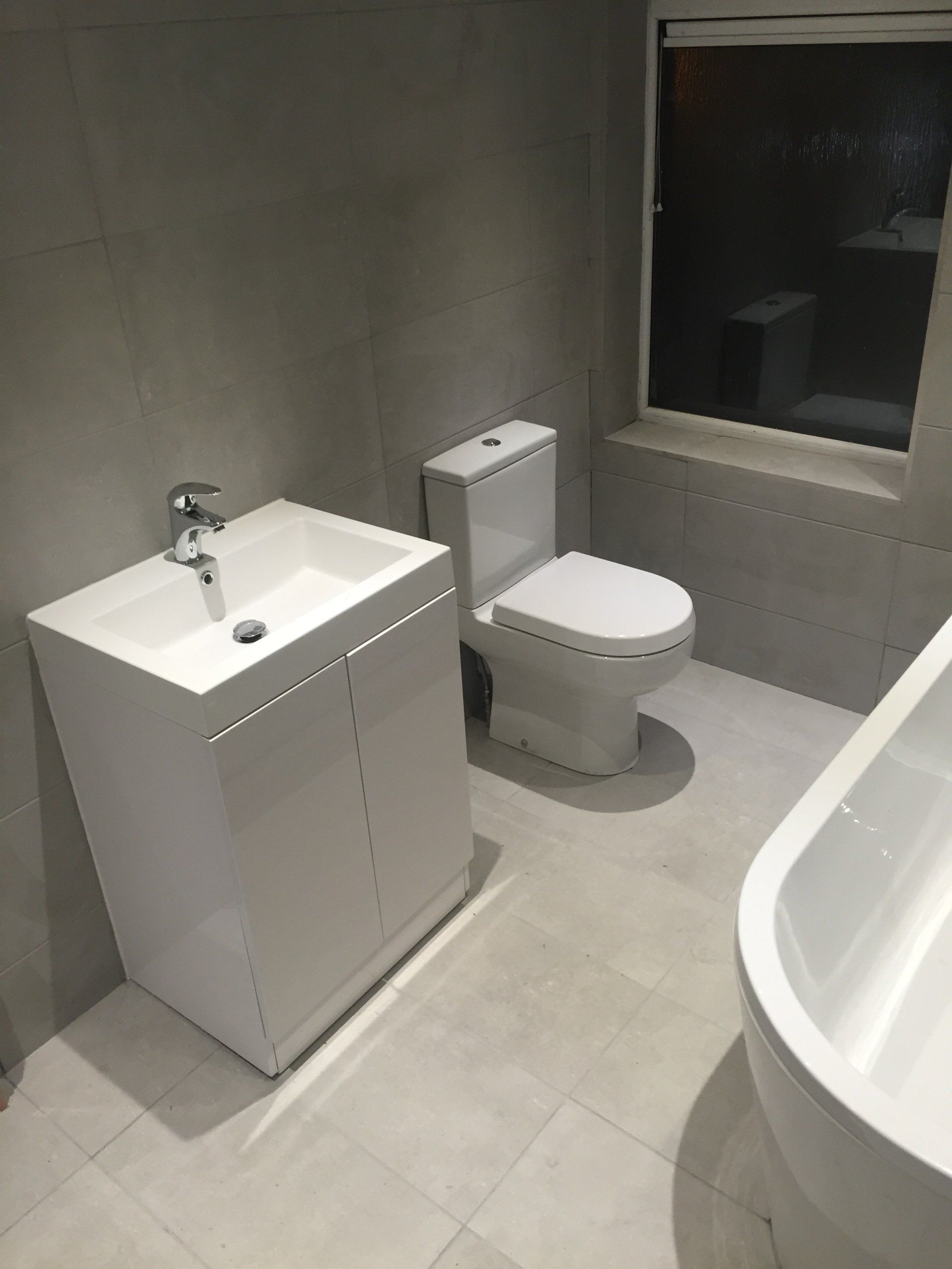 Modern bathroom with white sink cabinet, toilet, and bathtub; gray tile walls and floor.