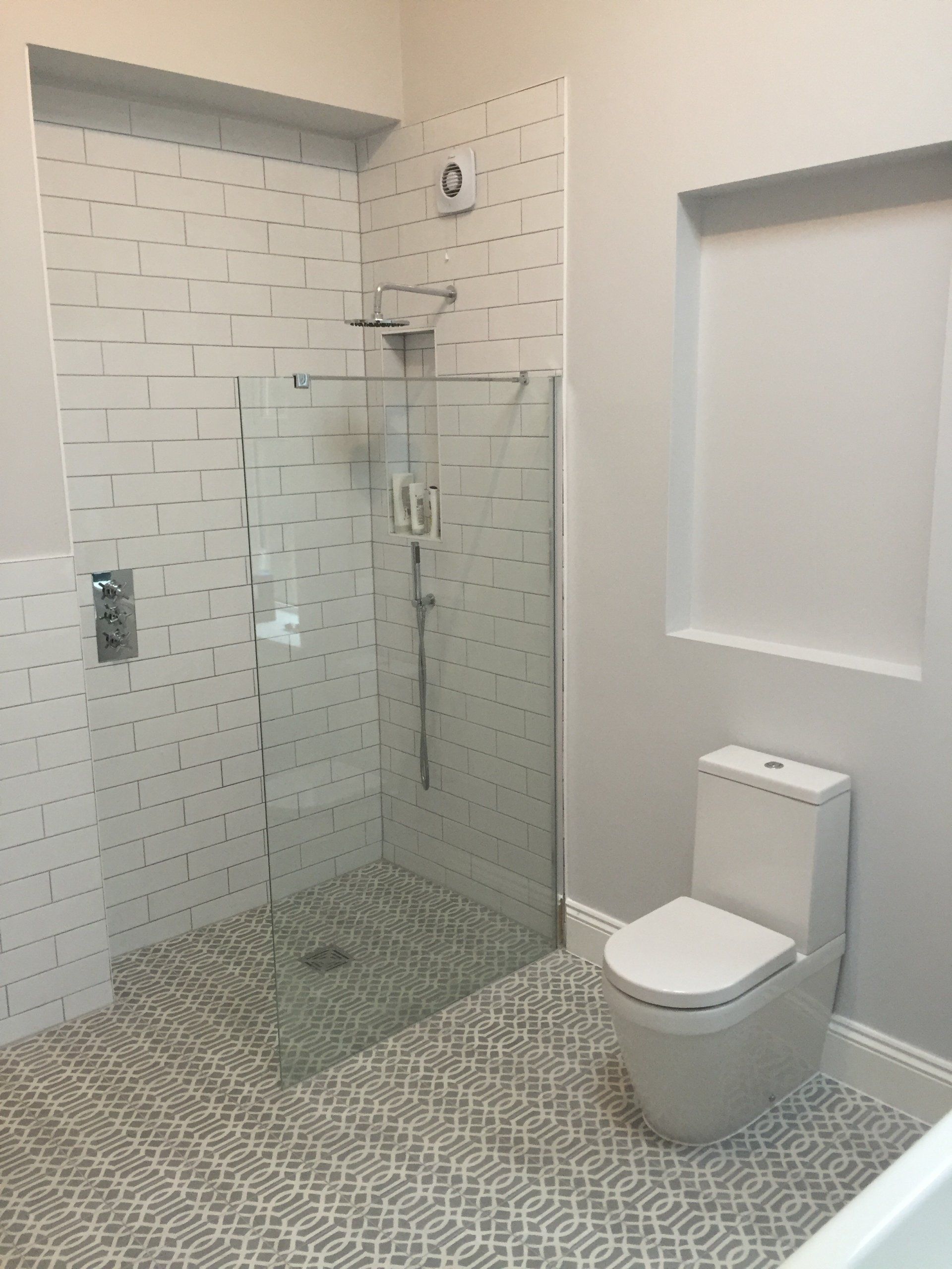 Modern bathroom with white subway tile shower, glass door, and patterned floor. A toilet sits beside a wall niche.