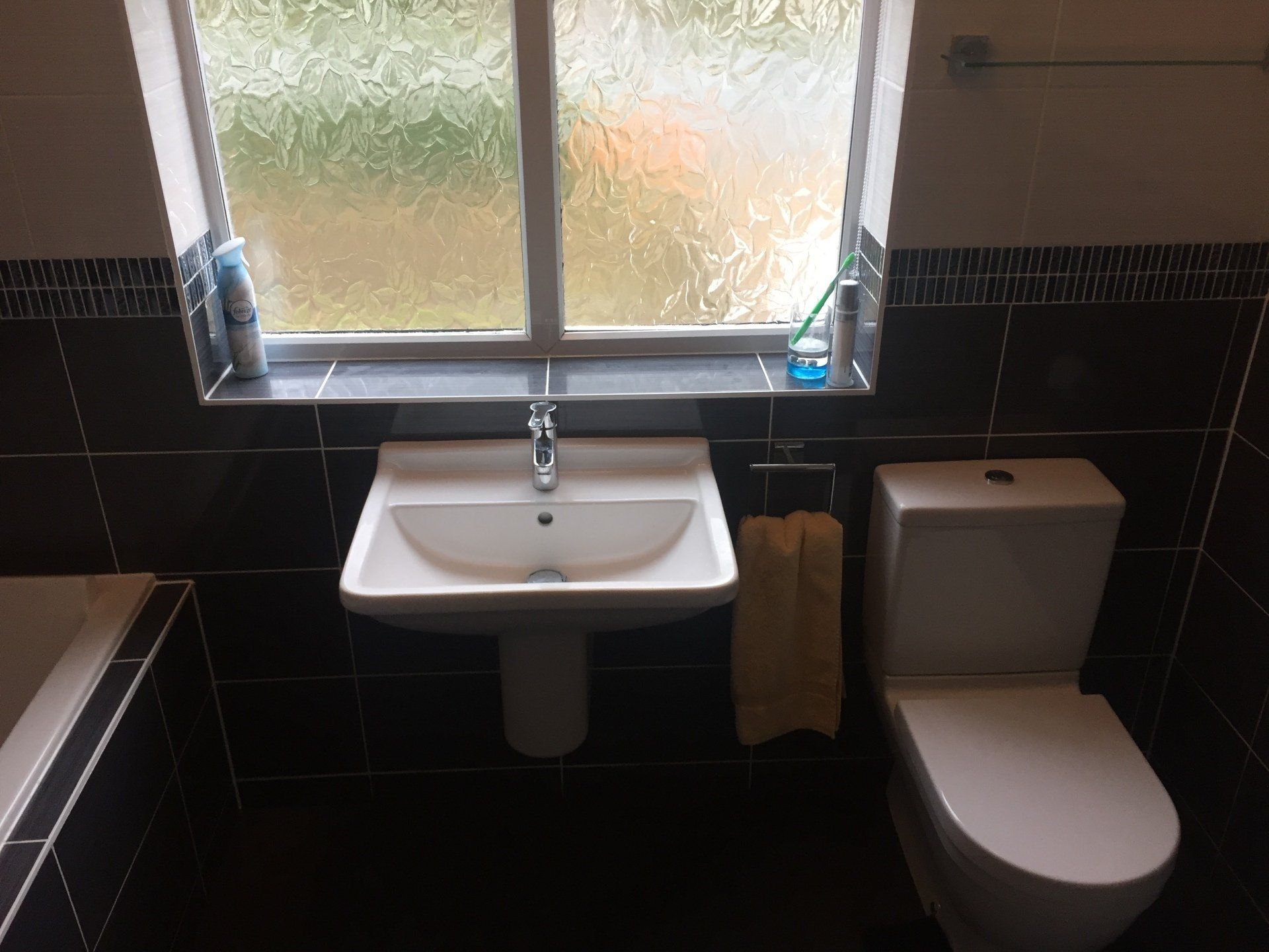 Bathroom with a white sink, toilet, and window. Black tile walls, a yellow towel, and toiletries on the window sill.