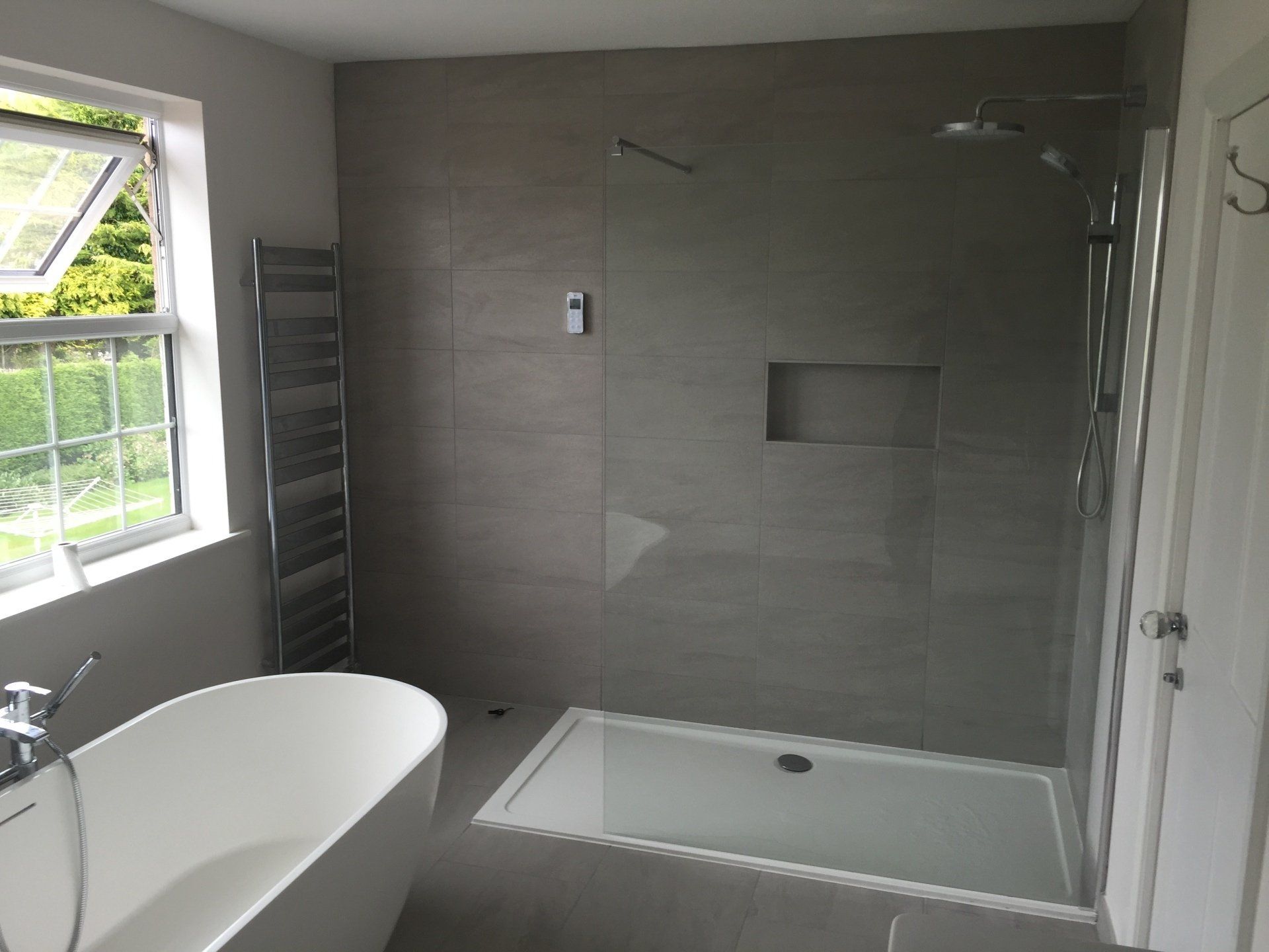 Modern bathroom with gray tiled shower, freestanding tub, window, and heated towel rack.