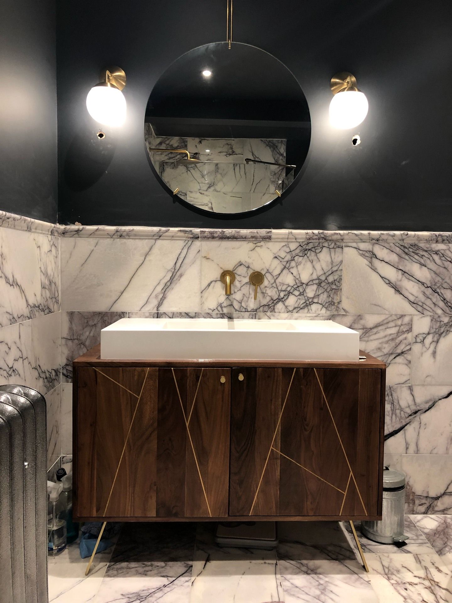 Bathroom with dark walls, marble tiles, wooden cabinet, round mirror, and brass fixtures.