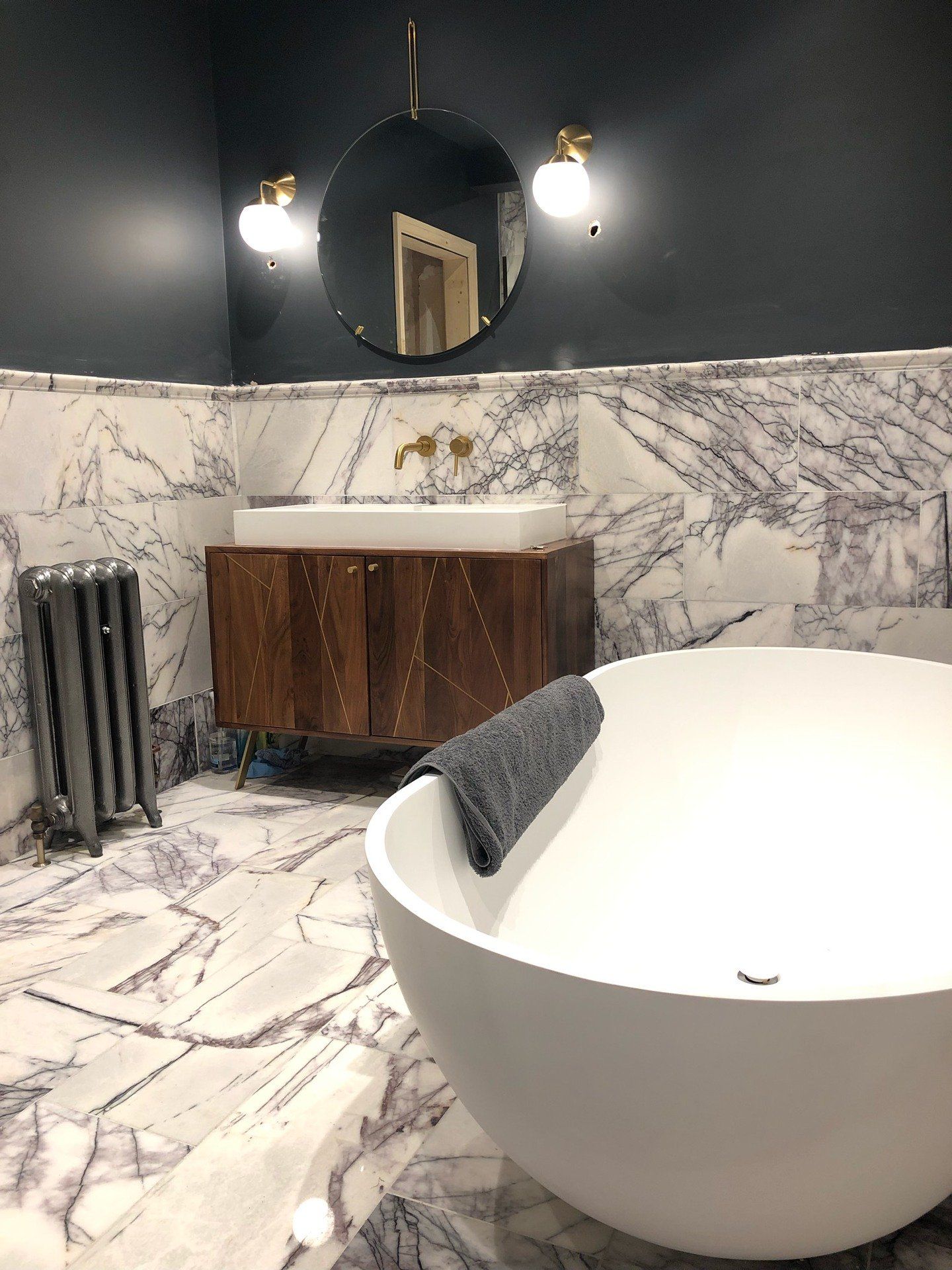Luxurious bathroom with marble tile, dark blue walls, a round mirror, and a freestanding tub.