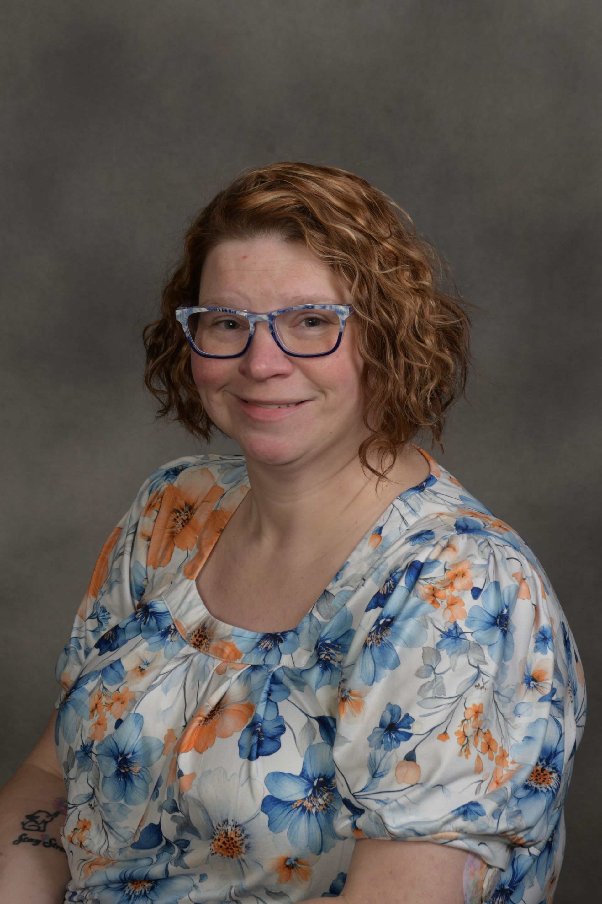 Woman wearing glasses, floral top, smiling, standing in front of a brick wall.