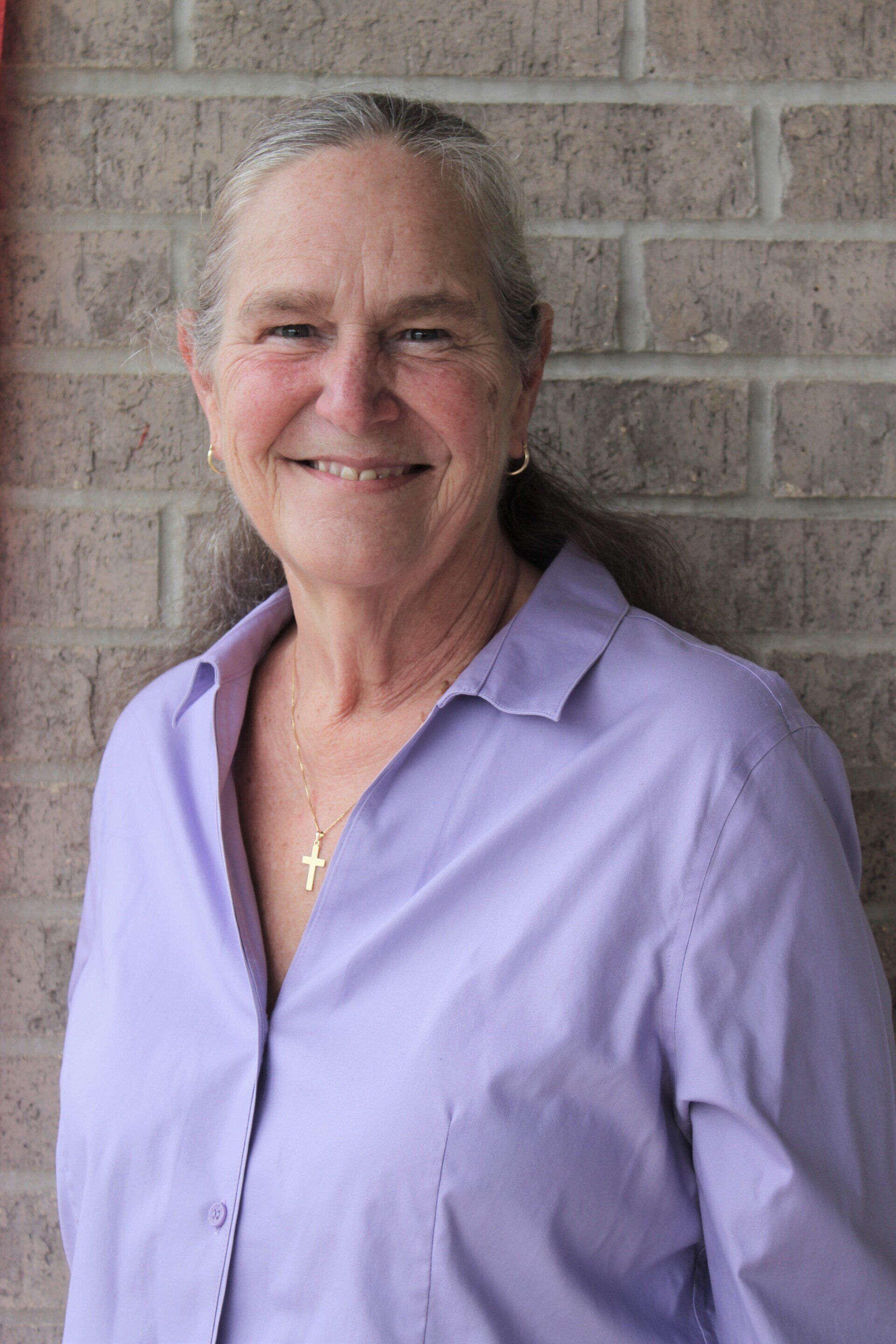 Anne Meyer is a white woman with salt-and-pepper hair pulled back in a ponytail. She is wearing a lavender shirt and a necklace with a gold cross pendant.