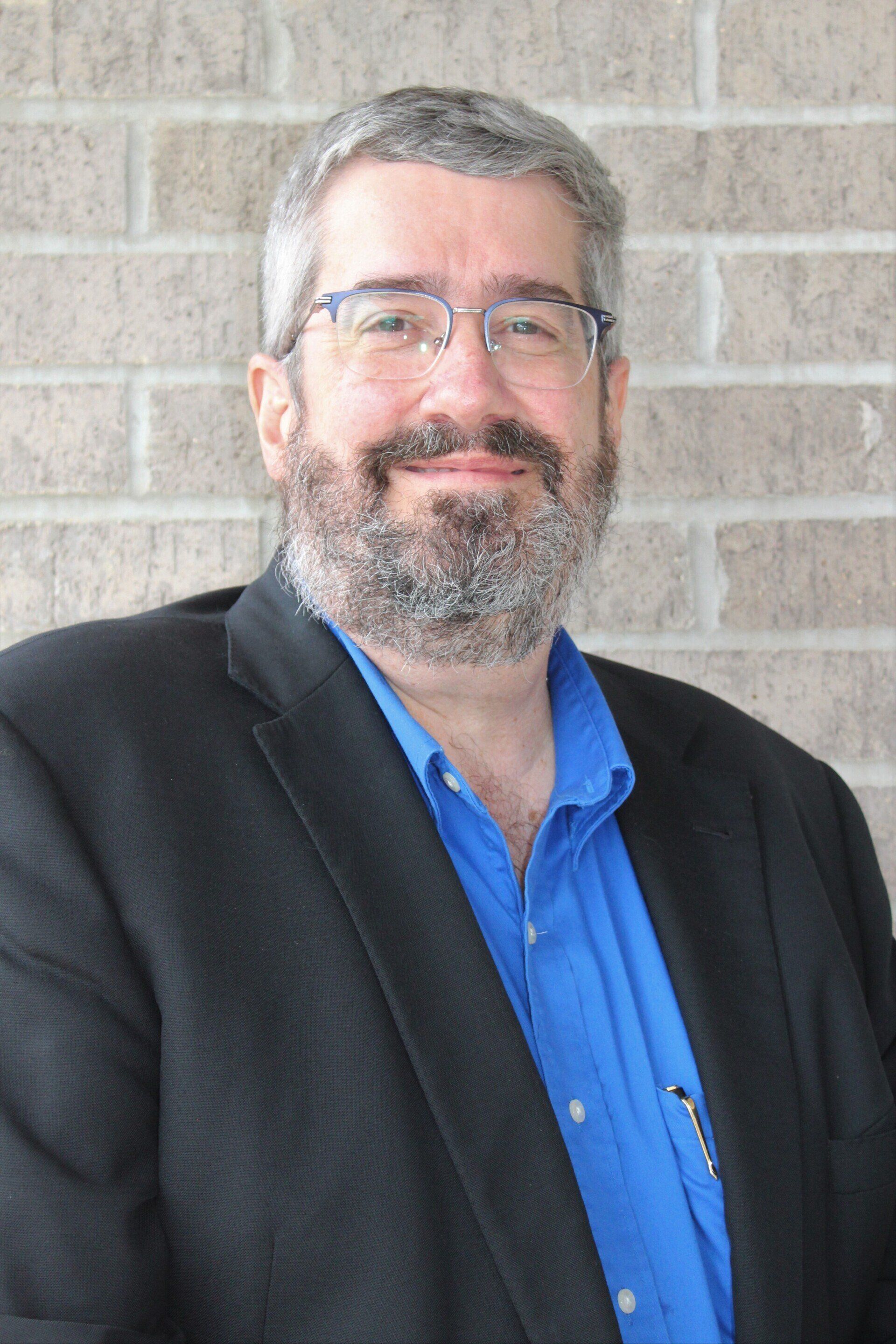 Lead Pastor Dan Dick, white man with salt-and-pepper hair and beard, wearing glasses and a blue shirt with black blazer