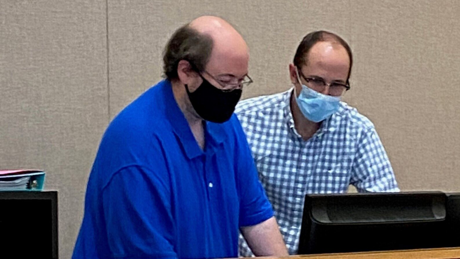 Two men wearing masks at a table, looking down at a screen. One in blue shirt, other in checkered shirt.