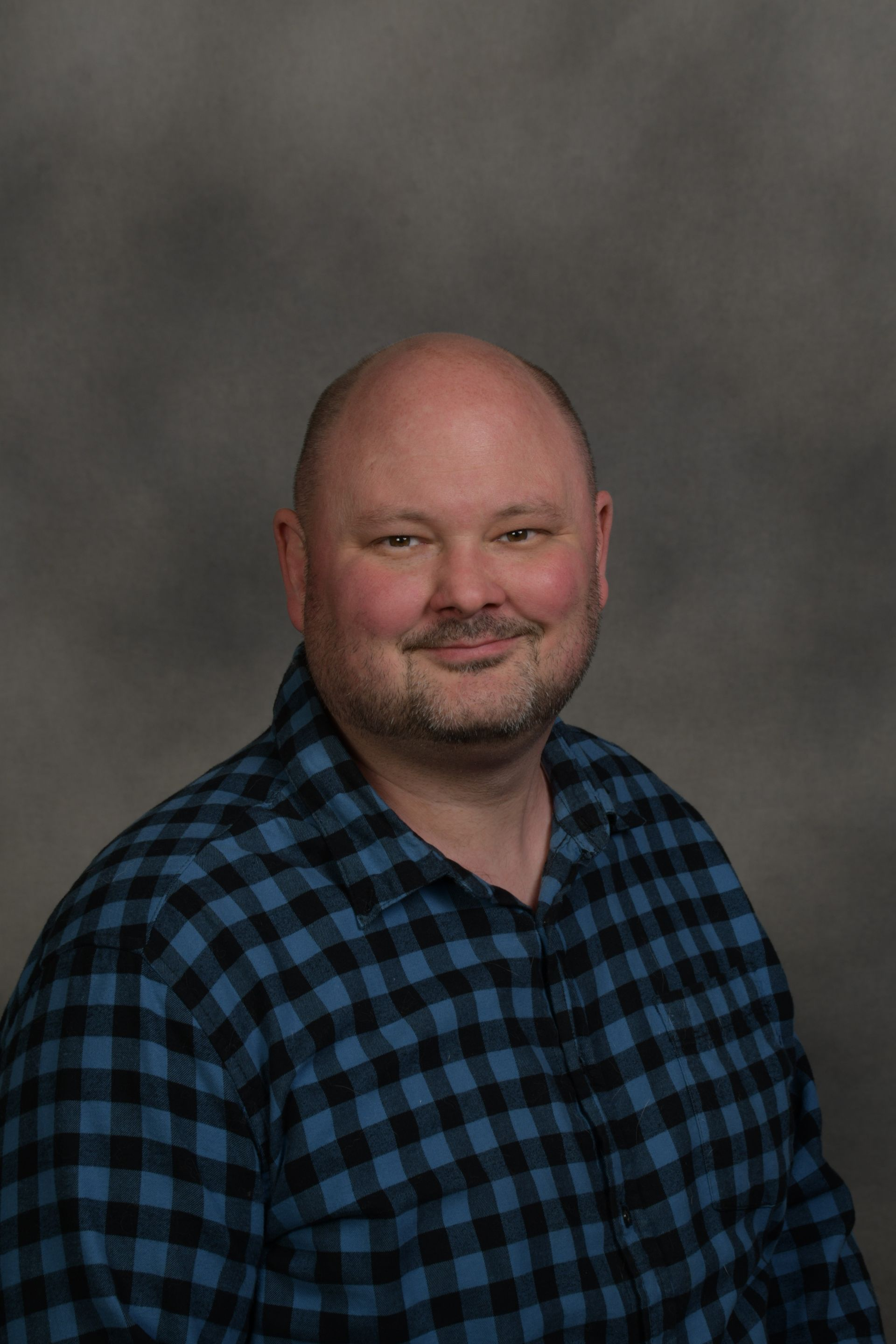 Man in tweed jacket smiles at camera, standing in front of a brick wall.