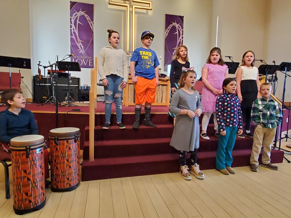 Children singing in Children's Choir. A child seated off to the left has a drum in front of him.