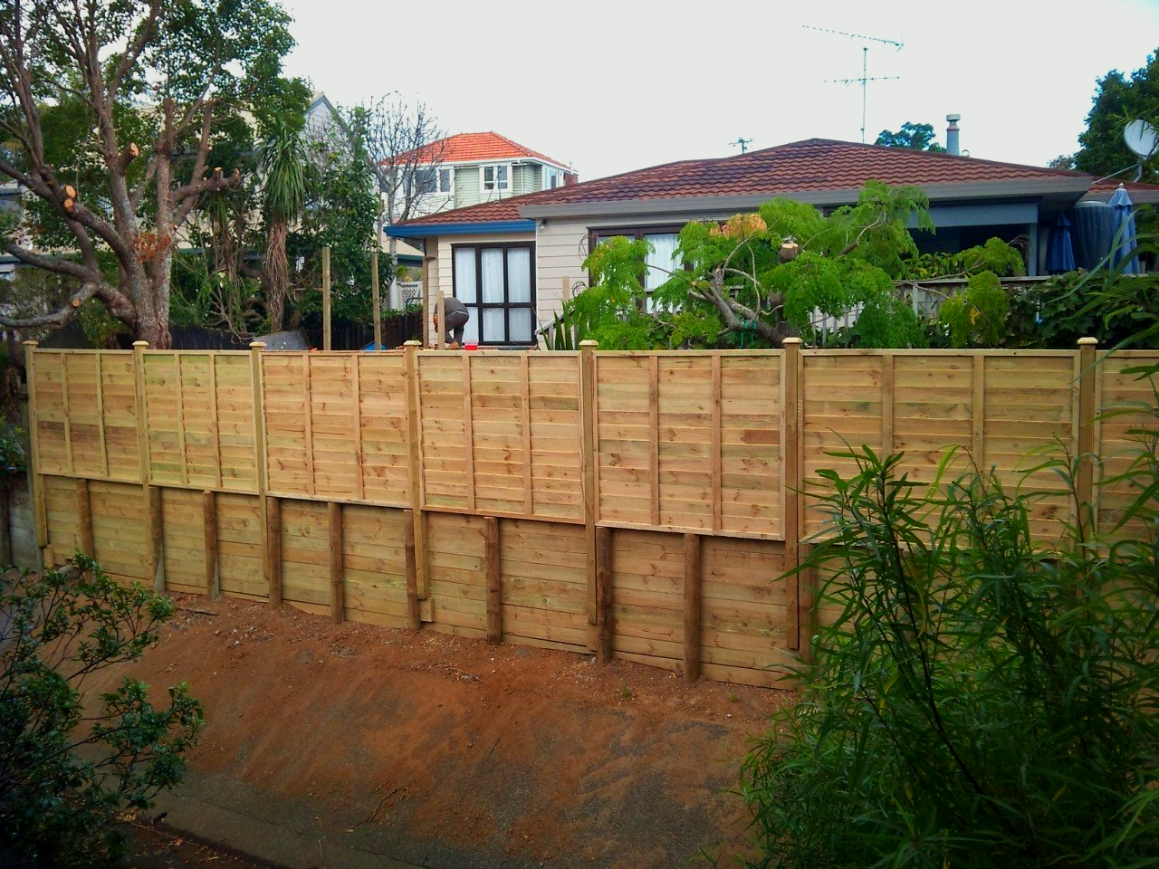 A wooden fence on top of a retaining wall, with a house in the background.