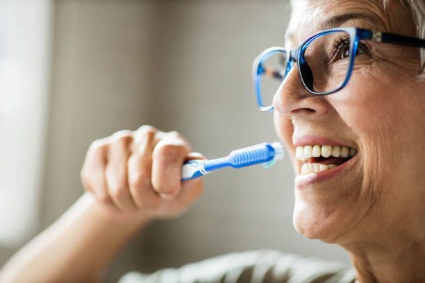 An elderly woman wearing glasses is brushing her teeth with a blue toothbrush.