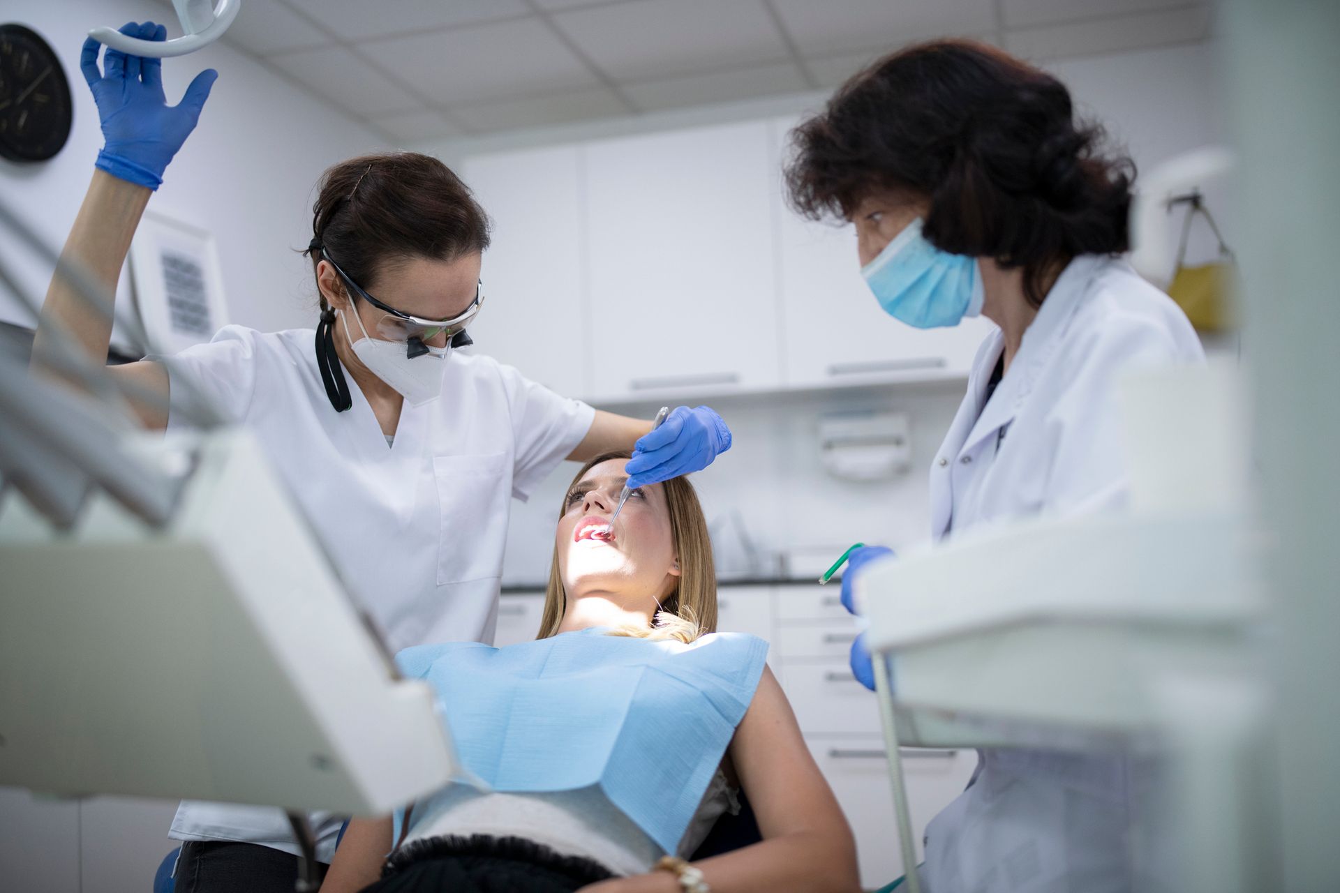 A woman is sitting in a dental chair getting her teeth examined by two dentists.