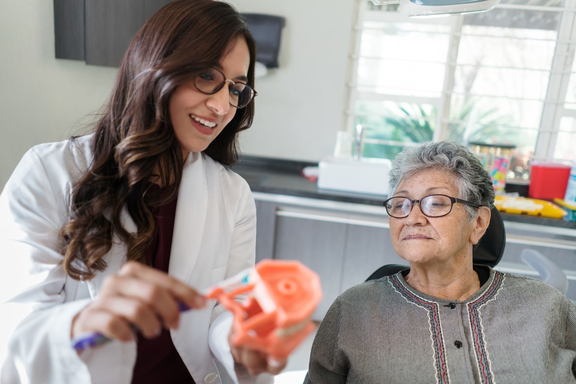 A dentist is showing an elderly woman how to brush her teeth.