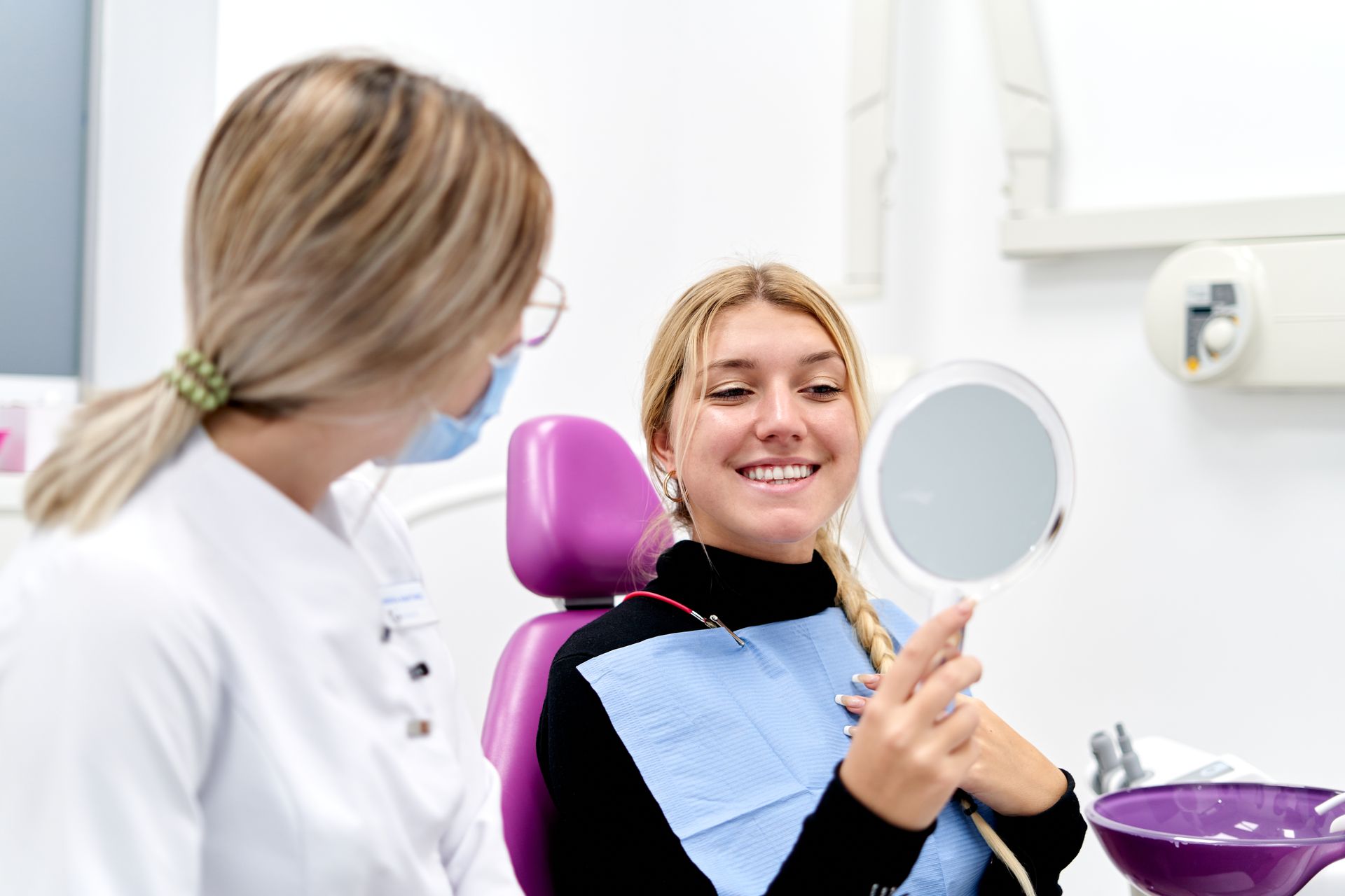 A blonde woman is sitting in a dental chair looking at her teeth in a mirror.