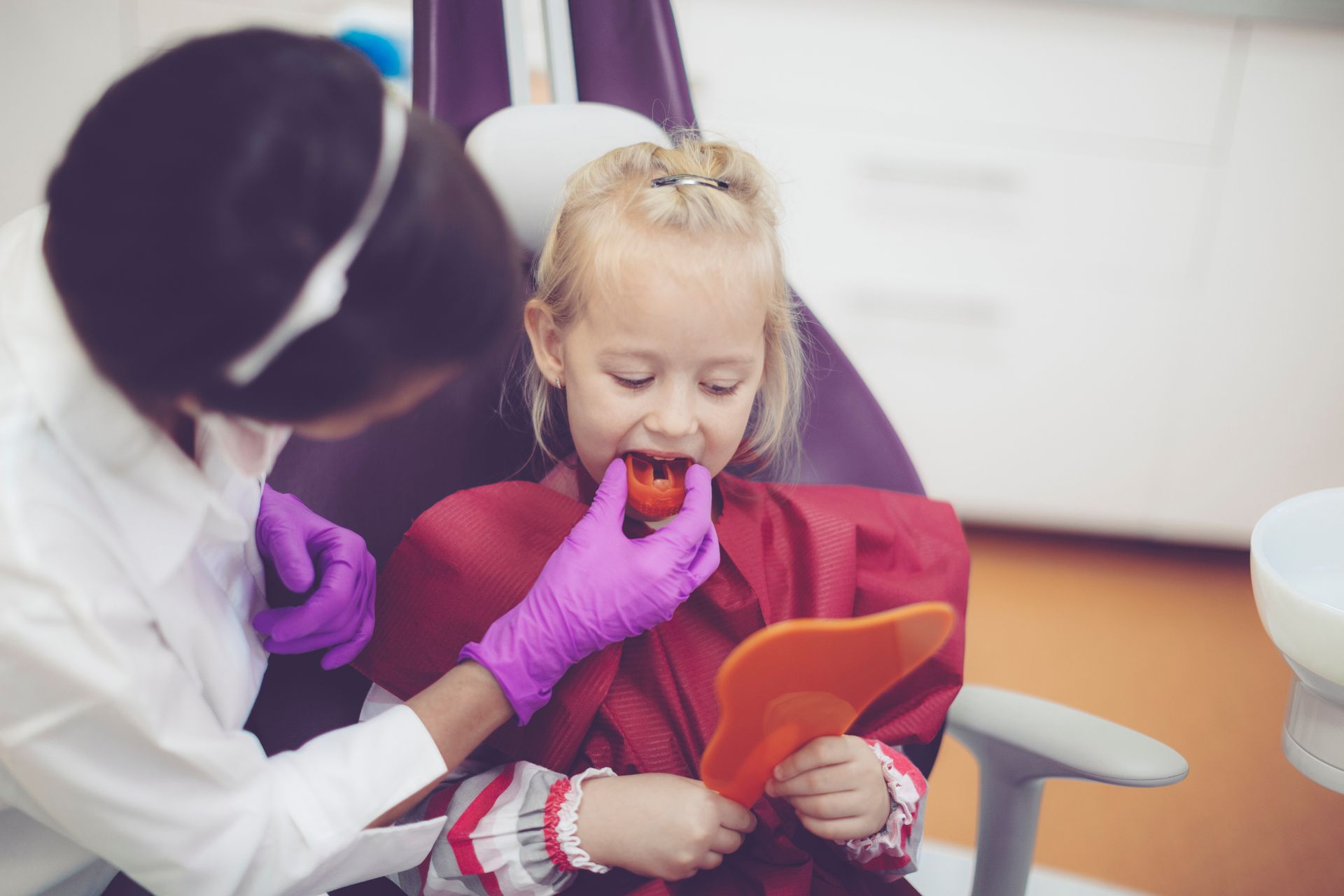 A little girl is sitting in a dental chair while a dentist examines her teeth.
