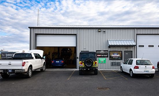 Three vehicles outside an auto repair shop, with a white truck, black SUV, and white sedan near open garage doors