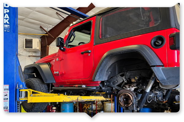 Red Jeep Wrangler lifted on a car hoist in an auto repair shop, wheels removed.