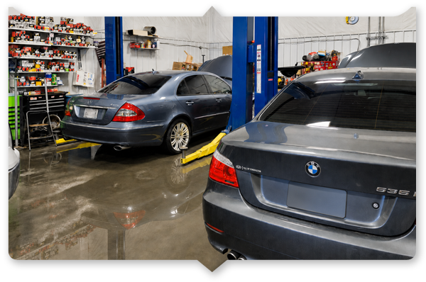 Two dark BMW sedans in a wet auto shop, one on a lift near shelves and tools.