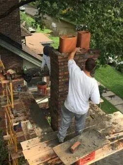 Worker on a rooftop laying bricks beside a chimney, with a canal and greenery below.