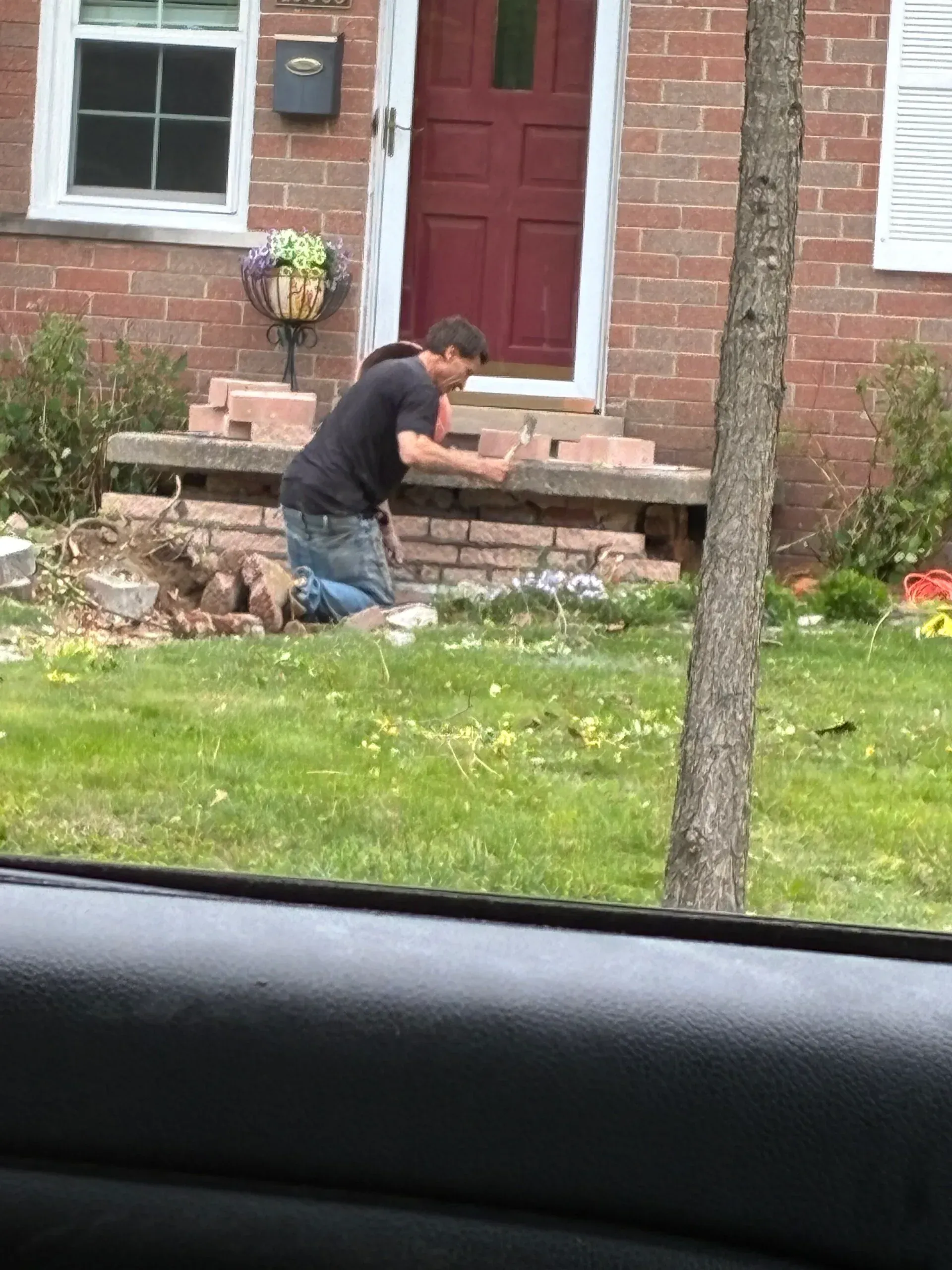 Person kneeling in a front yard beside a brick house with a red door