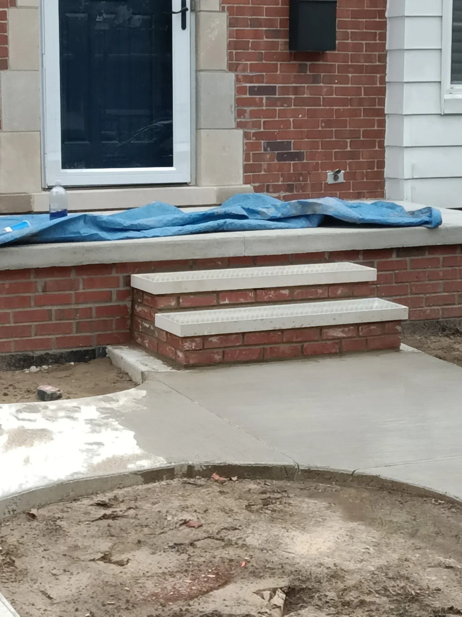Front steps of a brick house covered with a blue tarp, beside a concrete walkway.