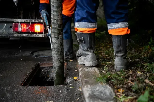 Worker in orange and blue uniform, boots, hose over a sewer opening, truck in background.