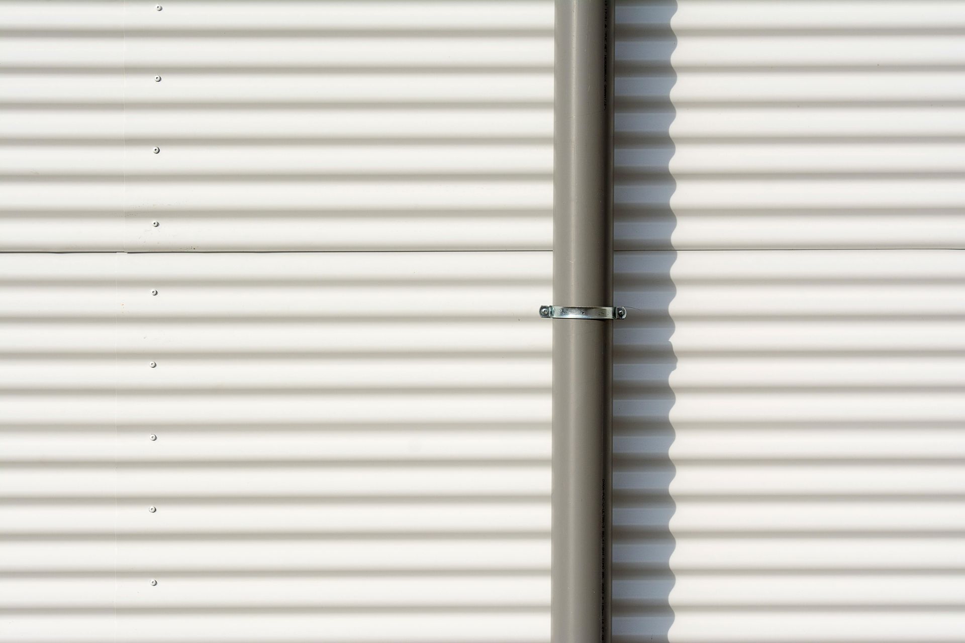 Gray pipe attached to a corrugated white wall with a metal clamp, casting a shadow.