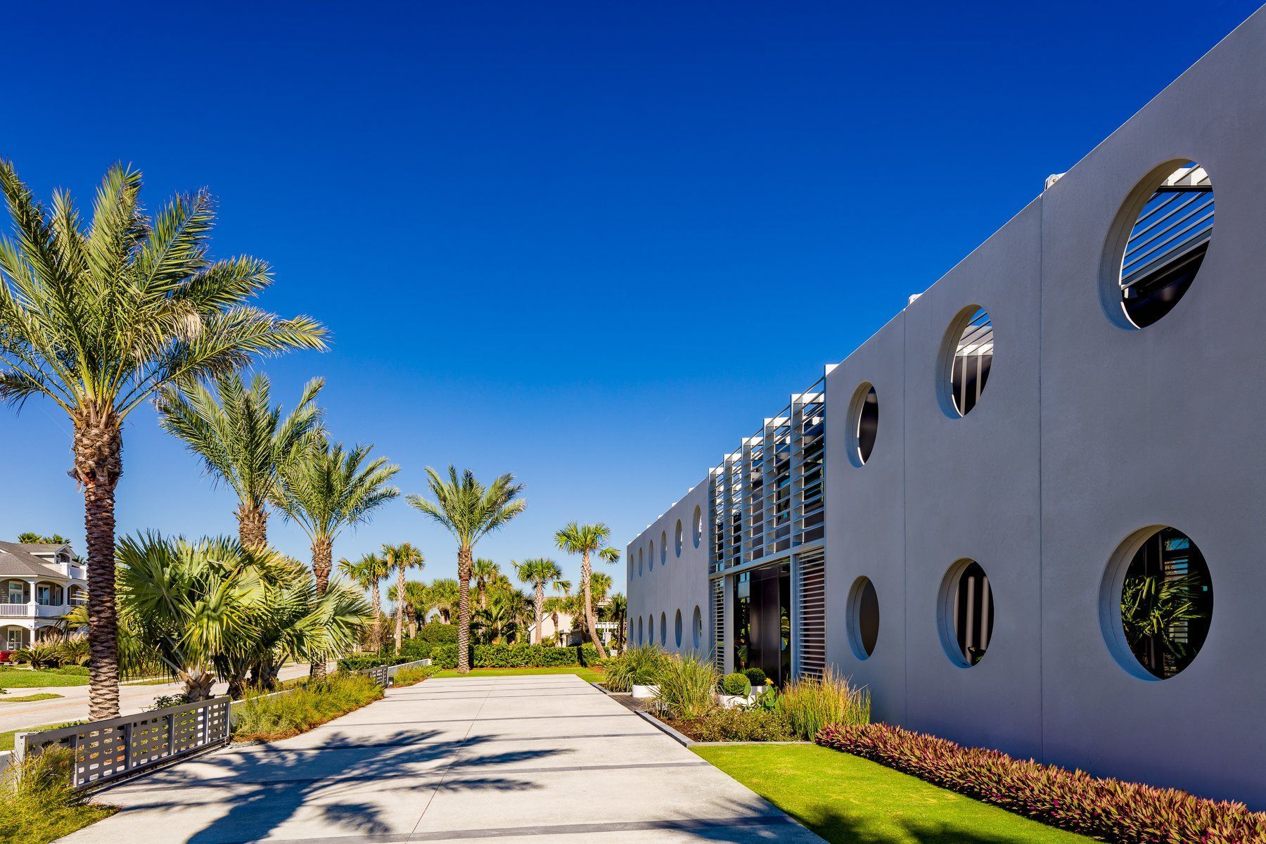 Exterior view of a modern building with round windows, palm trees, and a paved path under a blue sky.