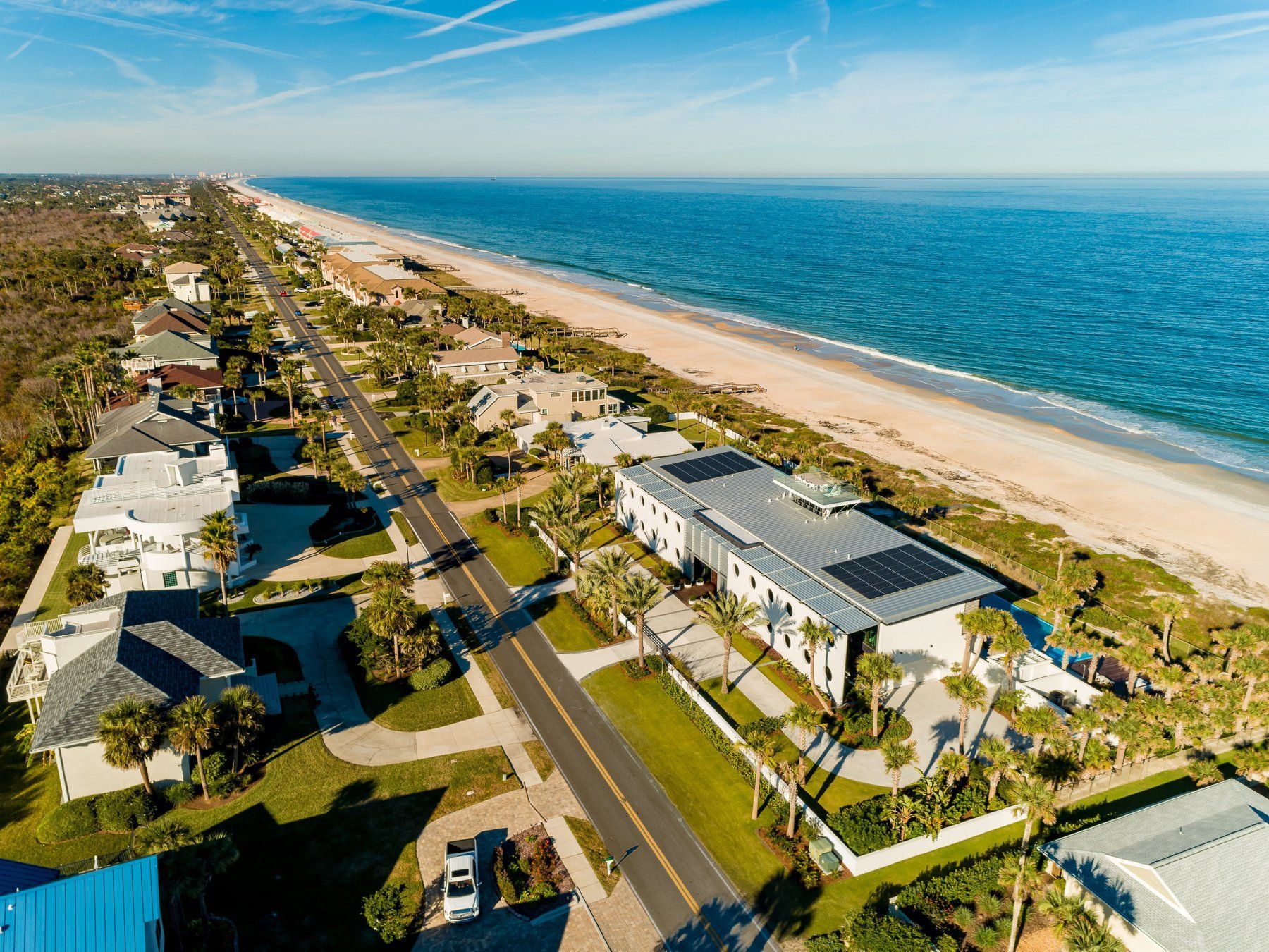 Aerial view of beach houses along a road, with the ocean on the right and blue sky overhead.