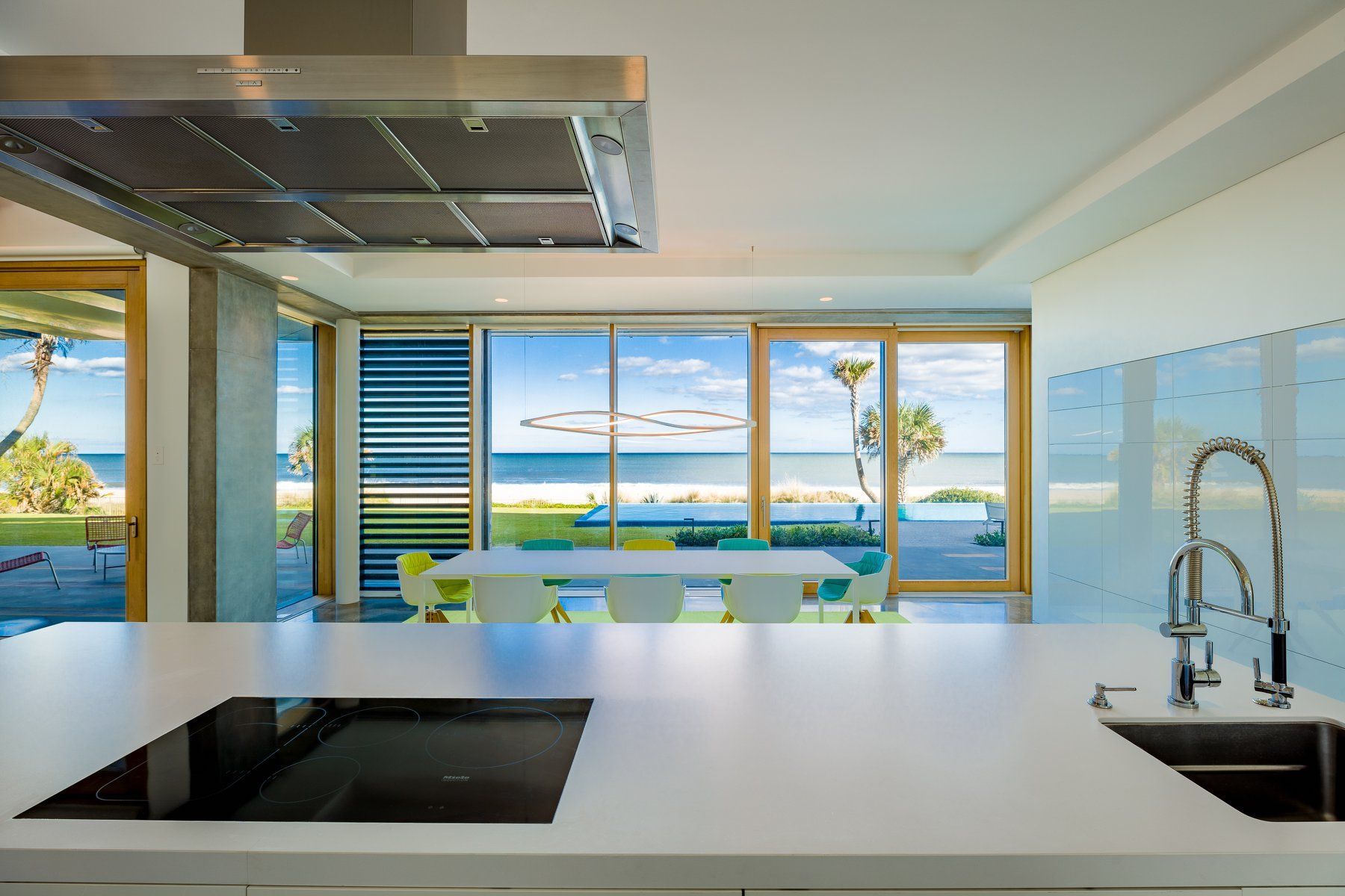 Modern kitchen with beach view: white counters, stainless steel hood, large windows, blue ocean, and a dining table.