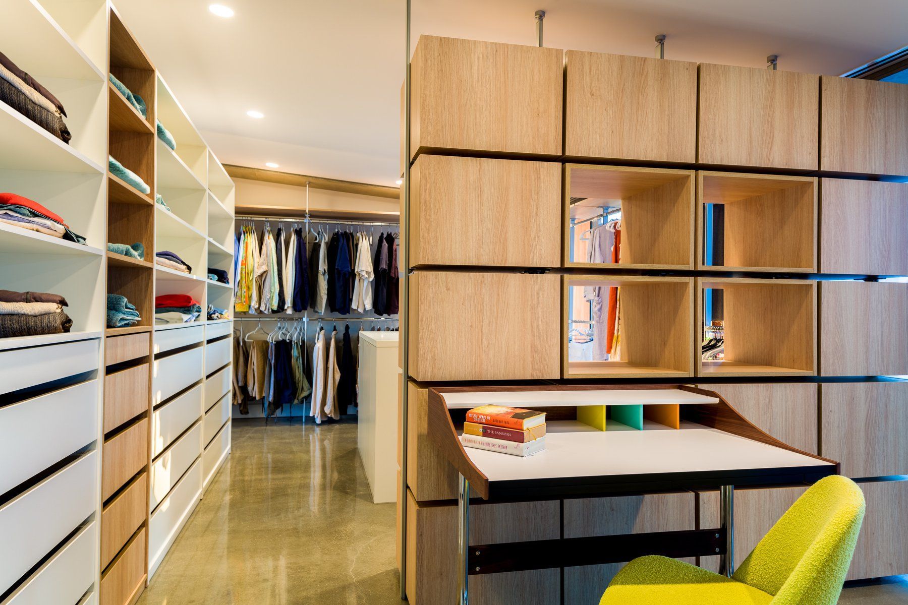 Modern closet with light wood, open shelves and drawers, built-in desk, clothes racks, and a yellow chair.