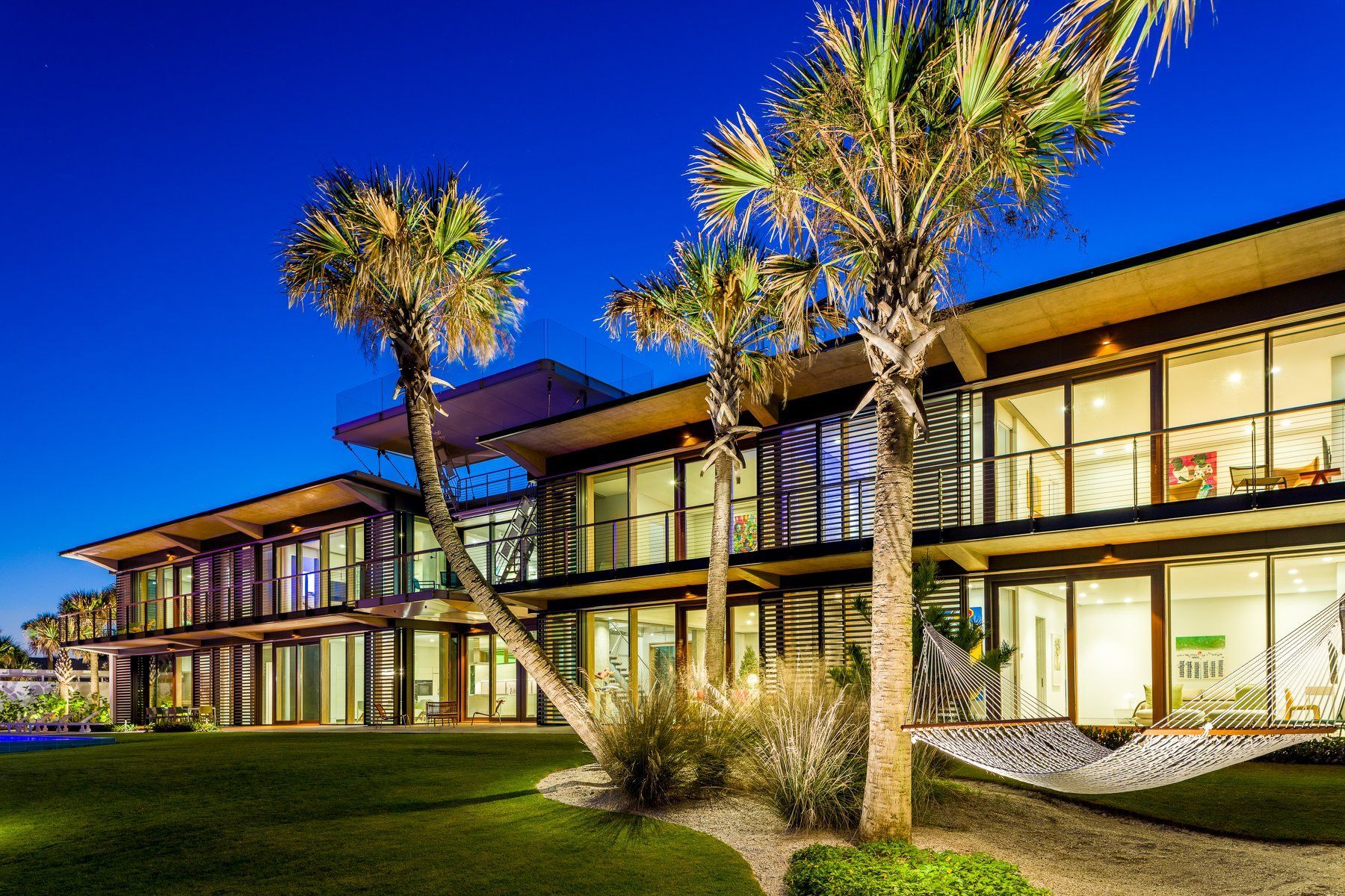 Modern beachfront condos at dusk; palm trees, grassy lawn, dark blue sky.