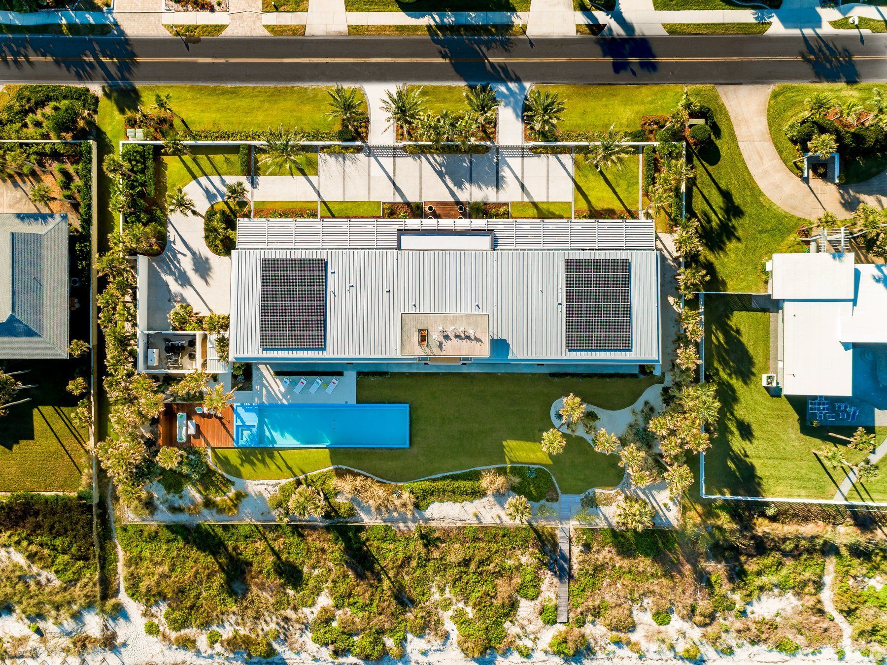 Overhead view of a modern, rectangular house with solar panels, a pool, and grassy lawn, on a beach with a road.