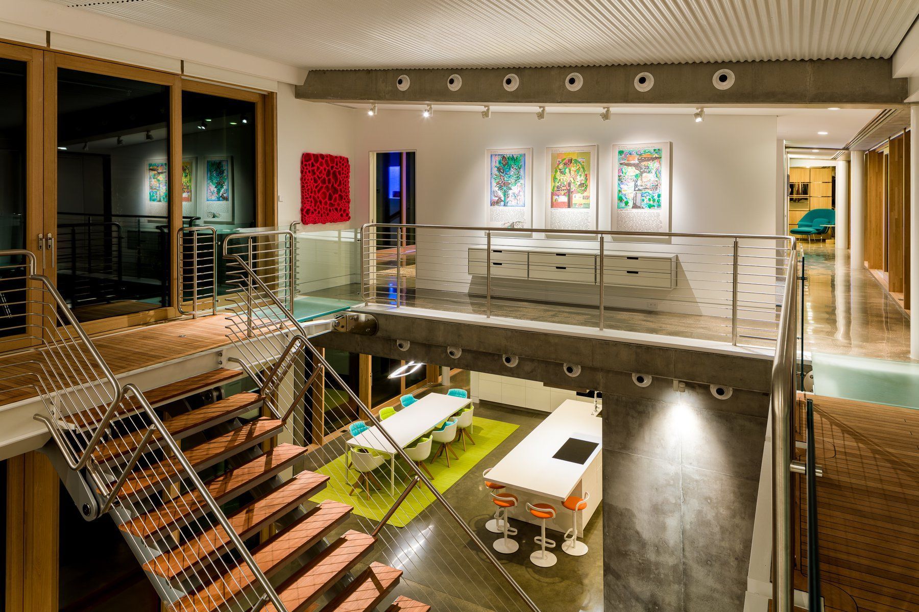Modern, two-story office space with stairs, gallery, workspace, and a kitchen. White, concrete, and glass with colorful artwork.