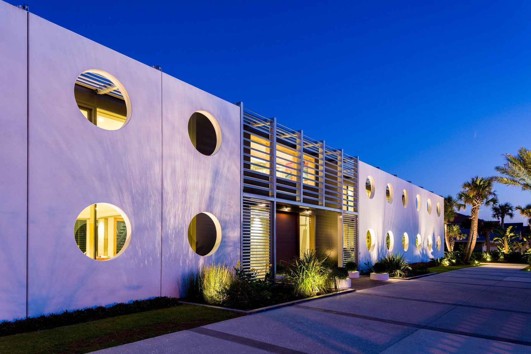 White building with circular windows, lit at dusk. A modern design with lush landscaping.