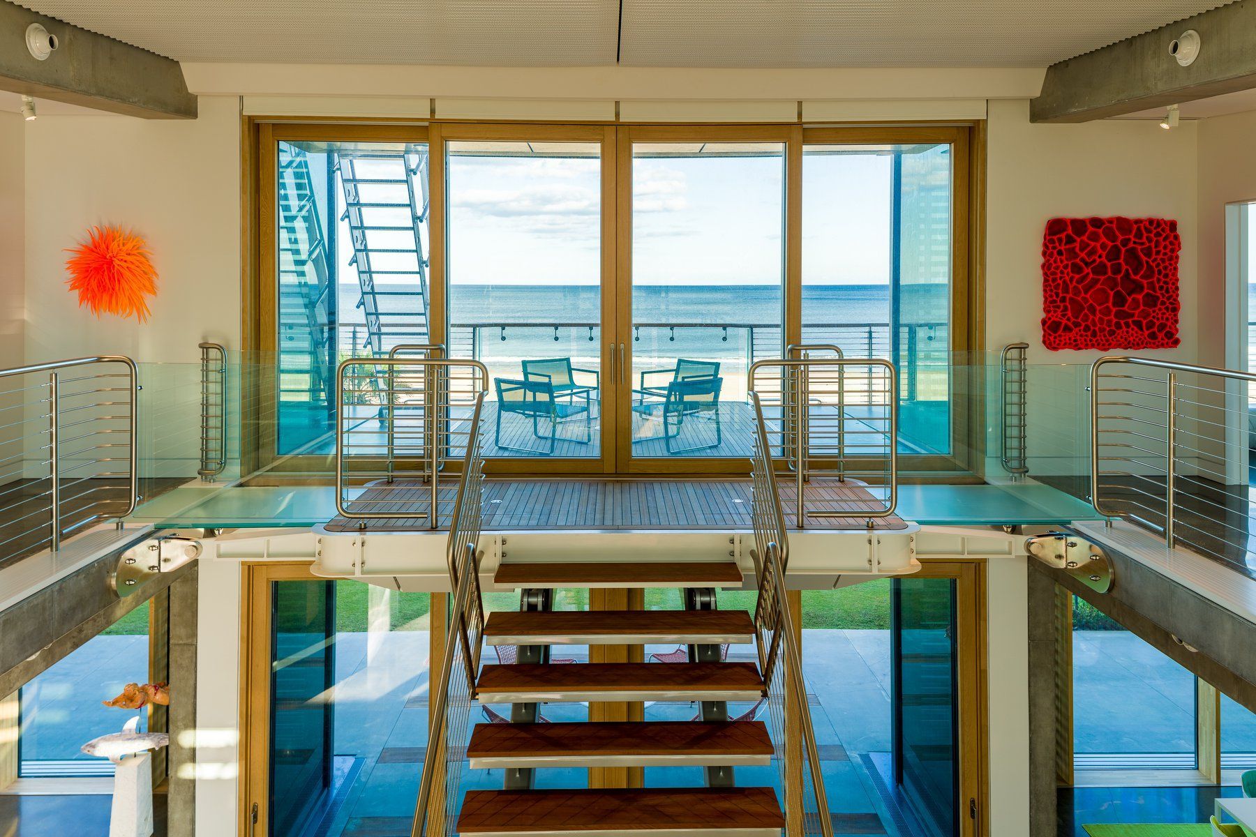 Interior view of a modern beach house with glass balcony, wooden stairs, and ocean view through large windows.