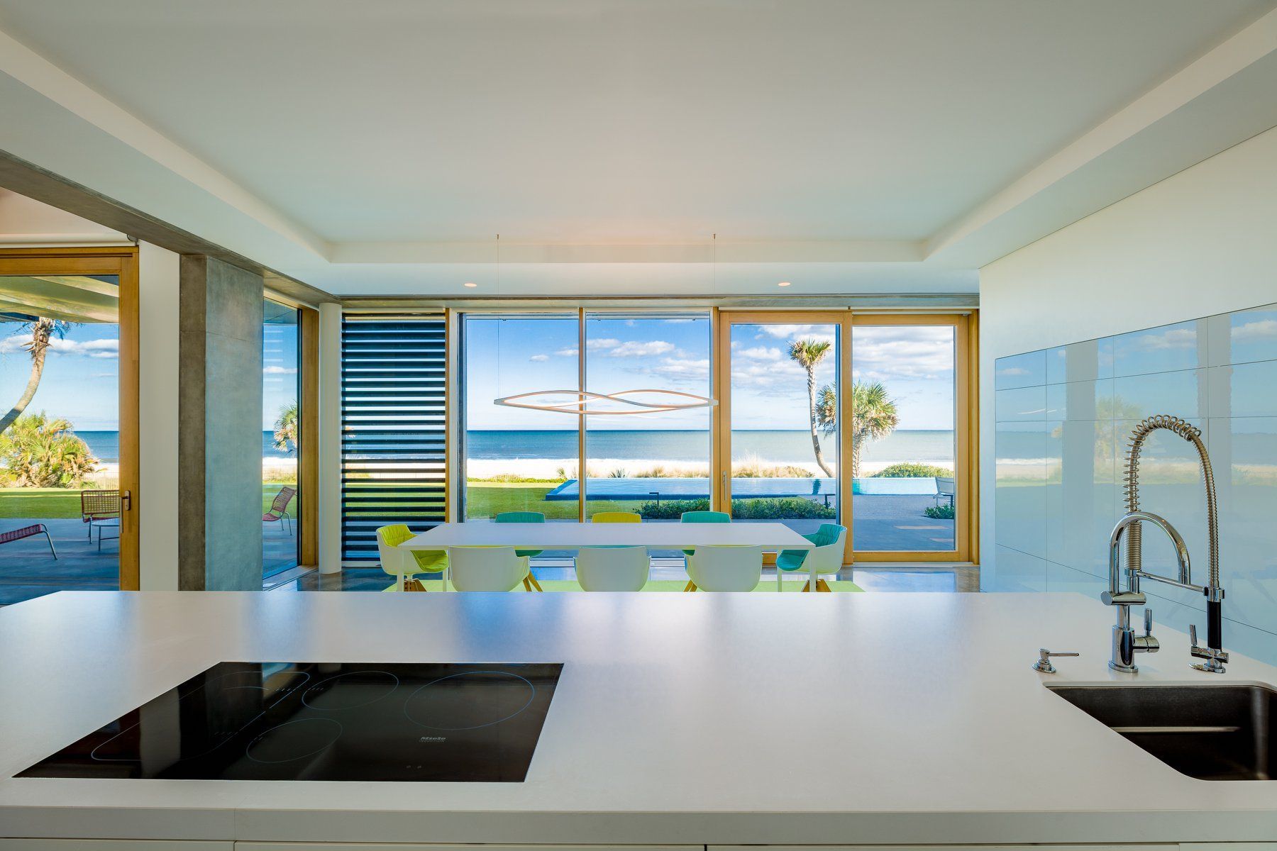 Bright kitchen with beach view: white countertop, blue-tiled wall, dining table with colorful chairs, and ocean through windows.
