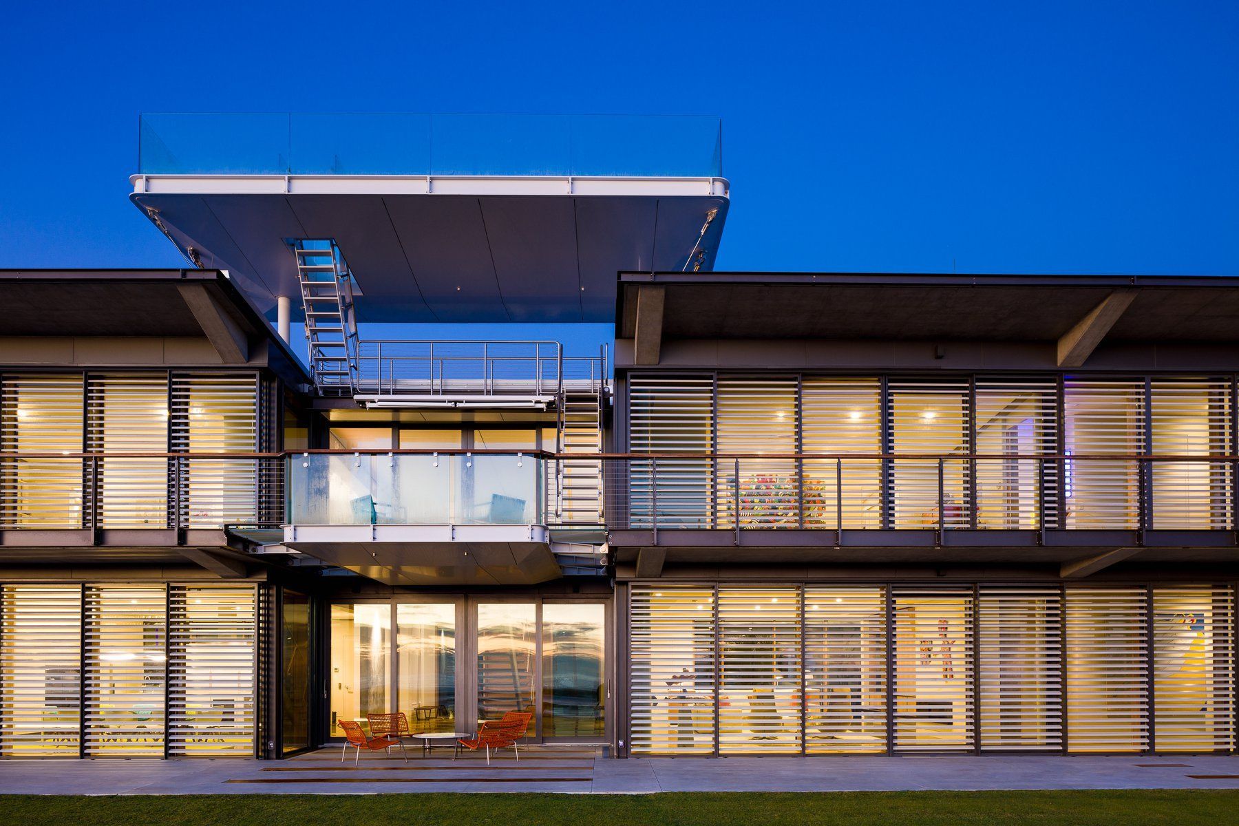 Modern two-story building with glass walls, a balcony, and a rooftop platform; dusk lighting.