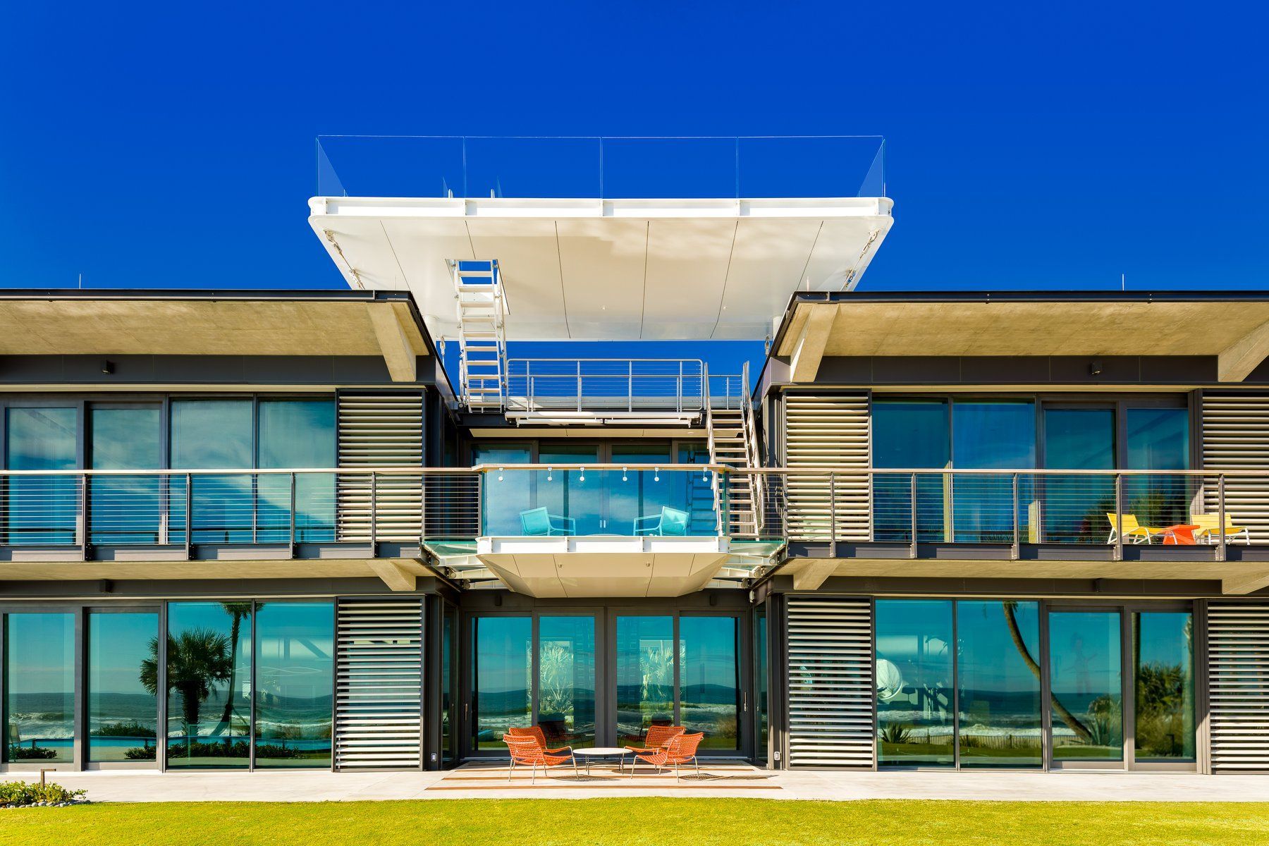 Modern beachfront home with glass walls, a rooftop deck, and a bright blue sky.