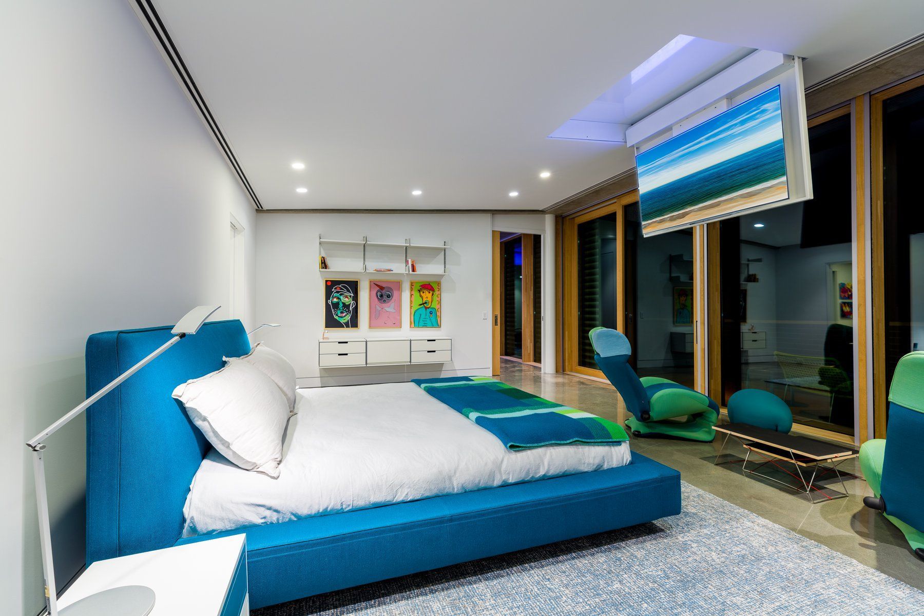 Bedroom with blue bed, ceiling-mounted TV, and seating by a glass door.