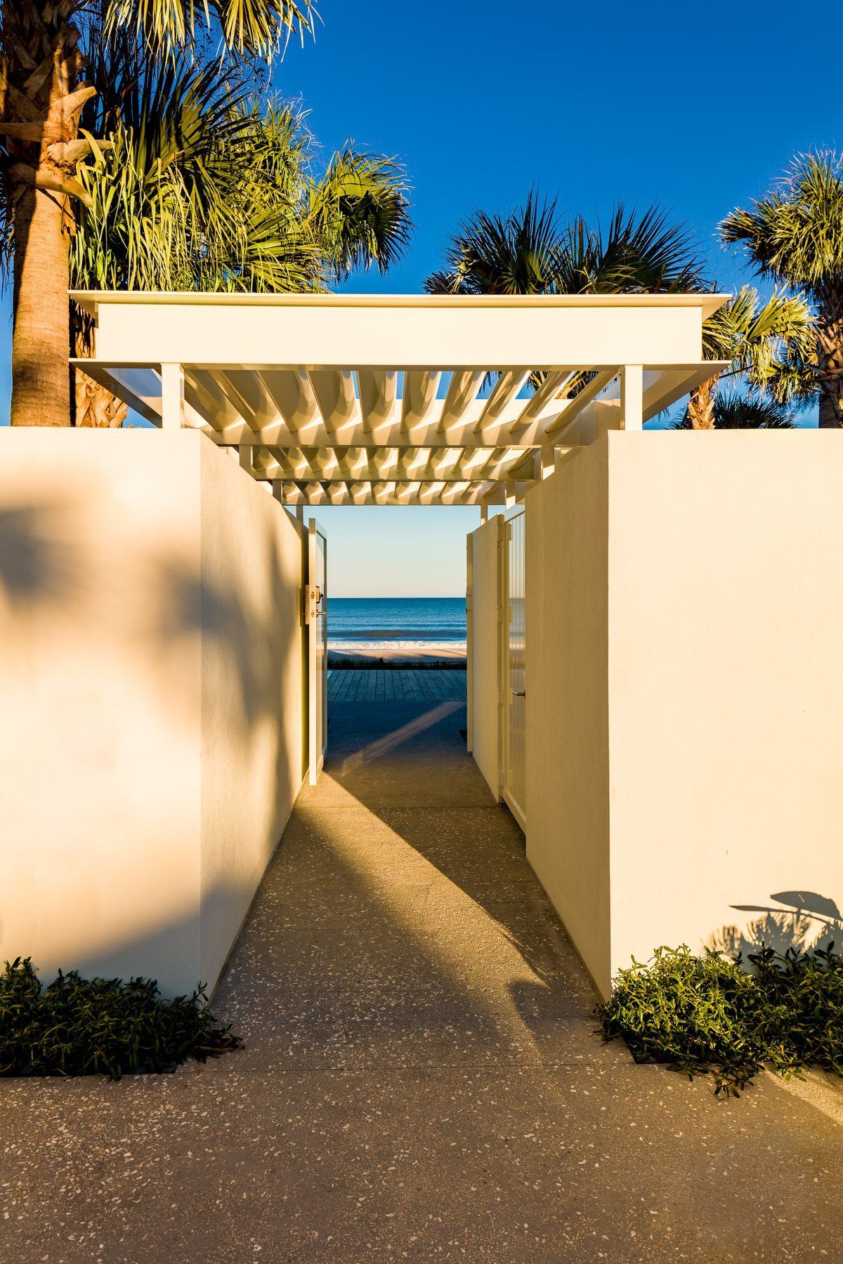 Passageway to the beach: white walls, overhead lattice, blue ocean visible. Sunlight casts long shadows.