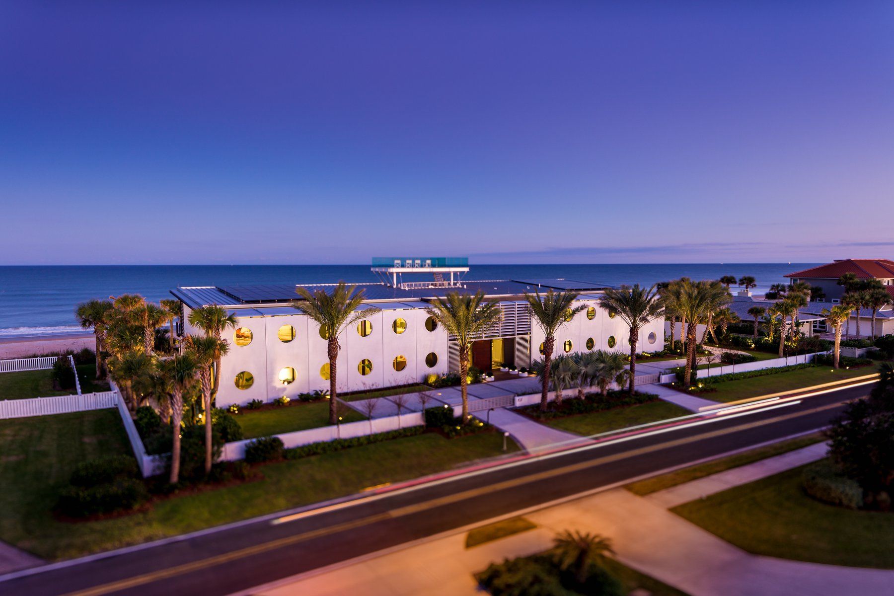Modern white beachfront home with round windows, palm trees, and a pier under a blue sky.