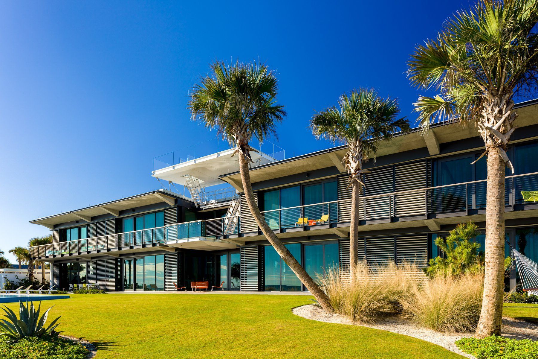 Modern beachfront home with palm trees, lush green lawn, and blue sky.
