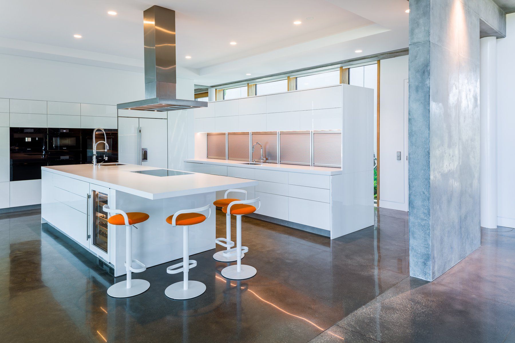 Modern white kitchen with island, orange stools, stainless steel accents, and a concrete floor.