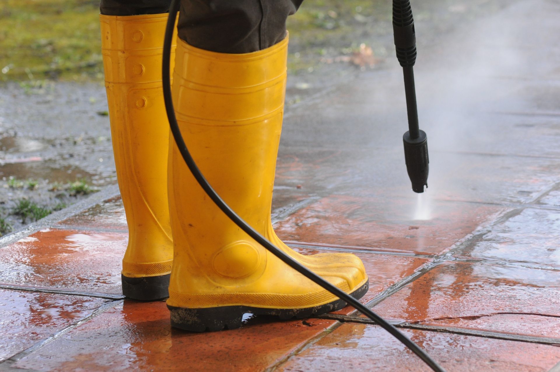 Person in yellow rain boots pressure-washing a wet pavement with a hose.