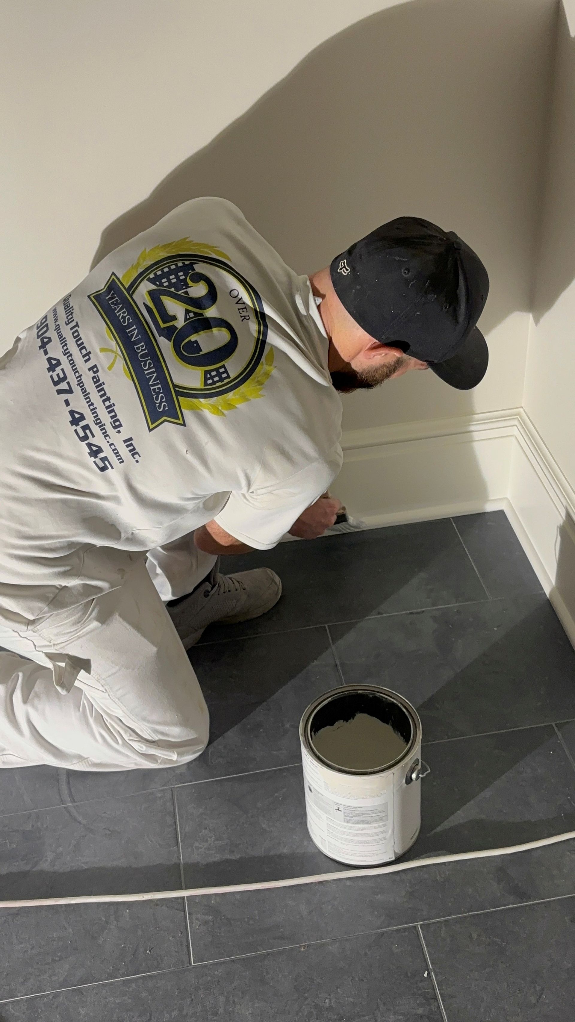 Painter kneeling to paint a baseboard in a room. He is wearing a hat and work clothes.