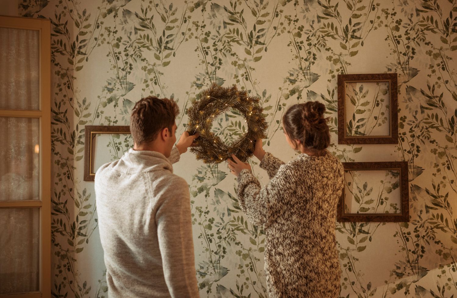 Couple hanging a wreath on a floral wallpapered wall, framed pictures, cozy setting.
